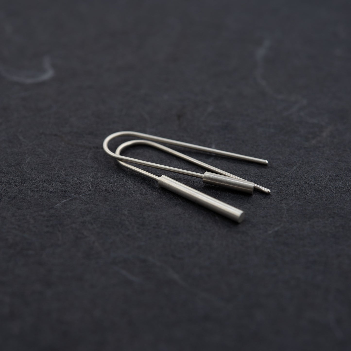 Artistic angled view of mismatched matte silver cylinder threader earrings, photographed on a black background – Paris jewelry in a clean, handmade style.