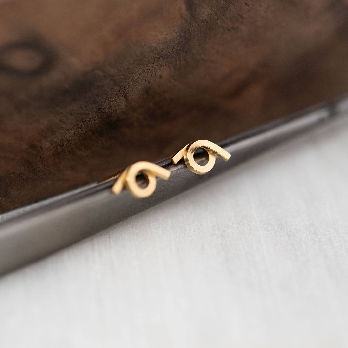 Vue rapprochée d'une paire de boucles d'oreilles en vermeil avec un design entrelacé, posées sur une surface en métal avec un fond texturé. Les boucles d'oreilles présentent une finition brillante, mettant en avant leur style moderne et élégant.