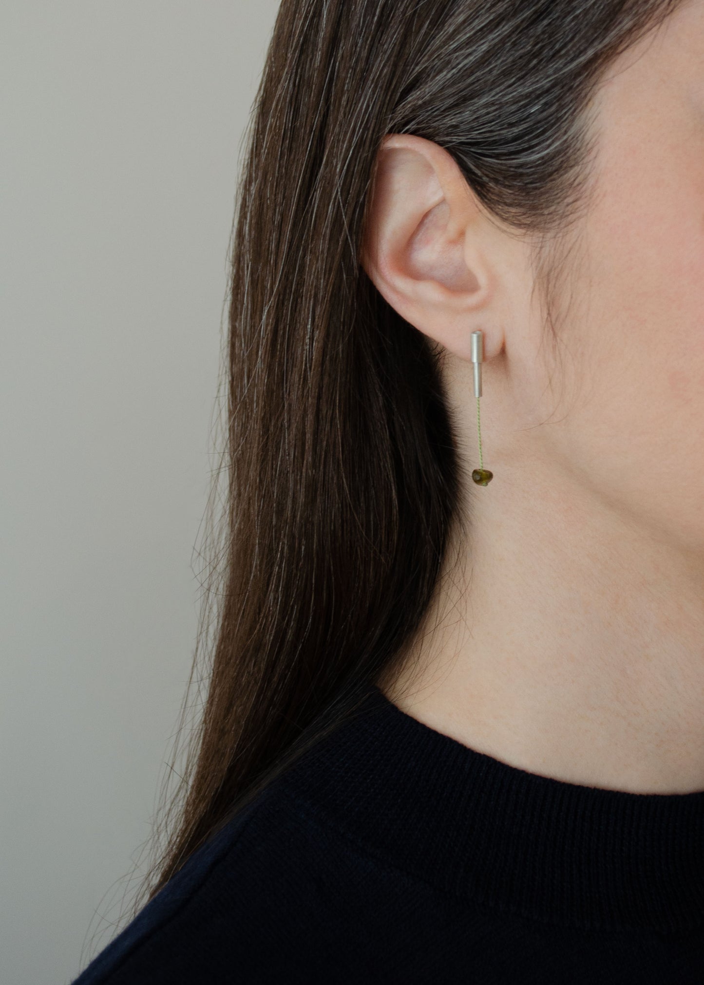 A half face of a woman wearing a pair of 37 millimeters length pendant earrings handmade in in Paris by A g J c from tube of matte sterling silver with a light green silk cord threaded through the center for hanging a green garnet stone.