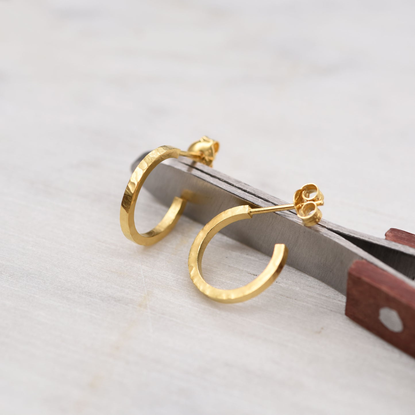 Vue rapprochée de boucles d'oreilles créoles en or martelé sur une surface blanche avec un outil gris en arrière-plan.