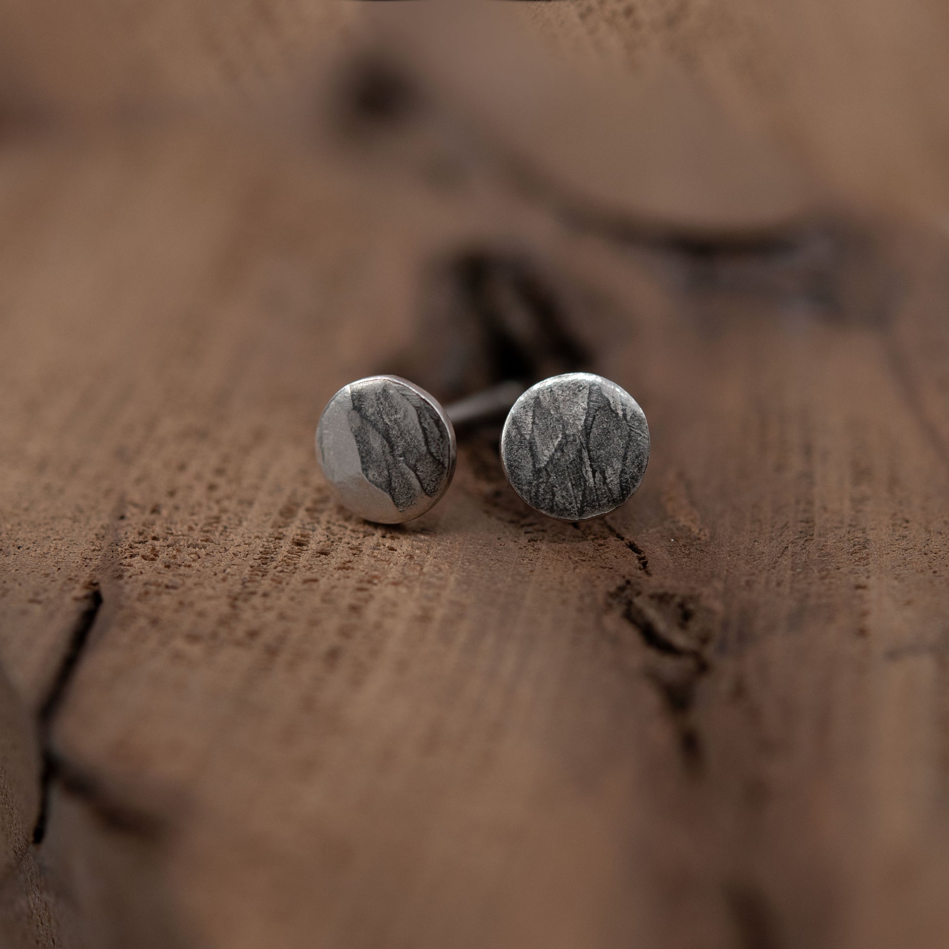 Close-up image of a pair of sterling silver stud earrings resting on a textured wooden surface. The earrings are round with a hammered finish, displaying a mix of shiny and matte areas that create a striking contrast. The natural tones of the wood provide a warm backdrop that highlights the elegant design of the silver earrings.