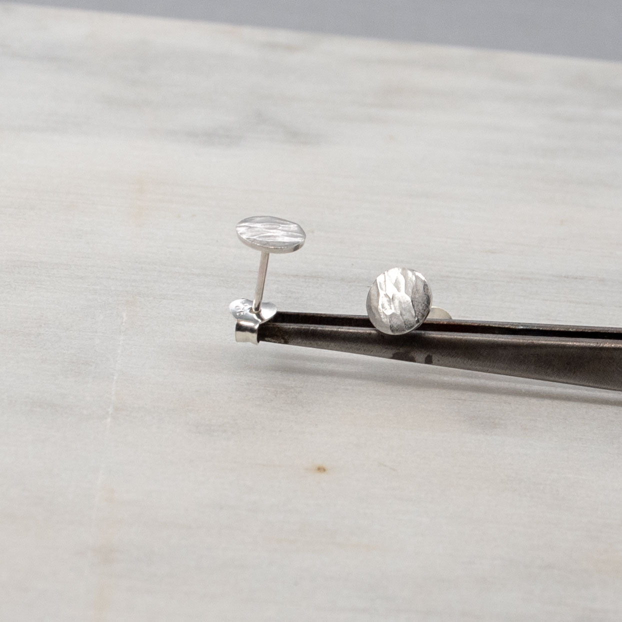 Image en gros plan de deux boucles d'oreilles en argent sterling posées sur une paire de pincettes. Les deux boucles d'oreilles présentent une finition texturée et martelée. L'arrière-plan est d'une couleur douce et neutre, mettant en valeur les boucles d'oreilles.