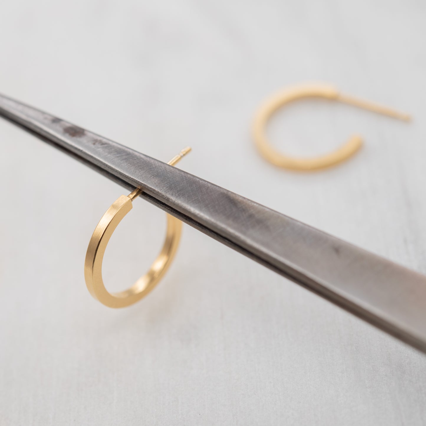 Paire de petites créoles dorées à l’or fin 24k photographiées sur un outil de bijoutier, fabrication artisanale parisienne.
