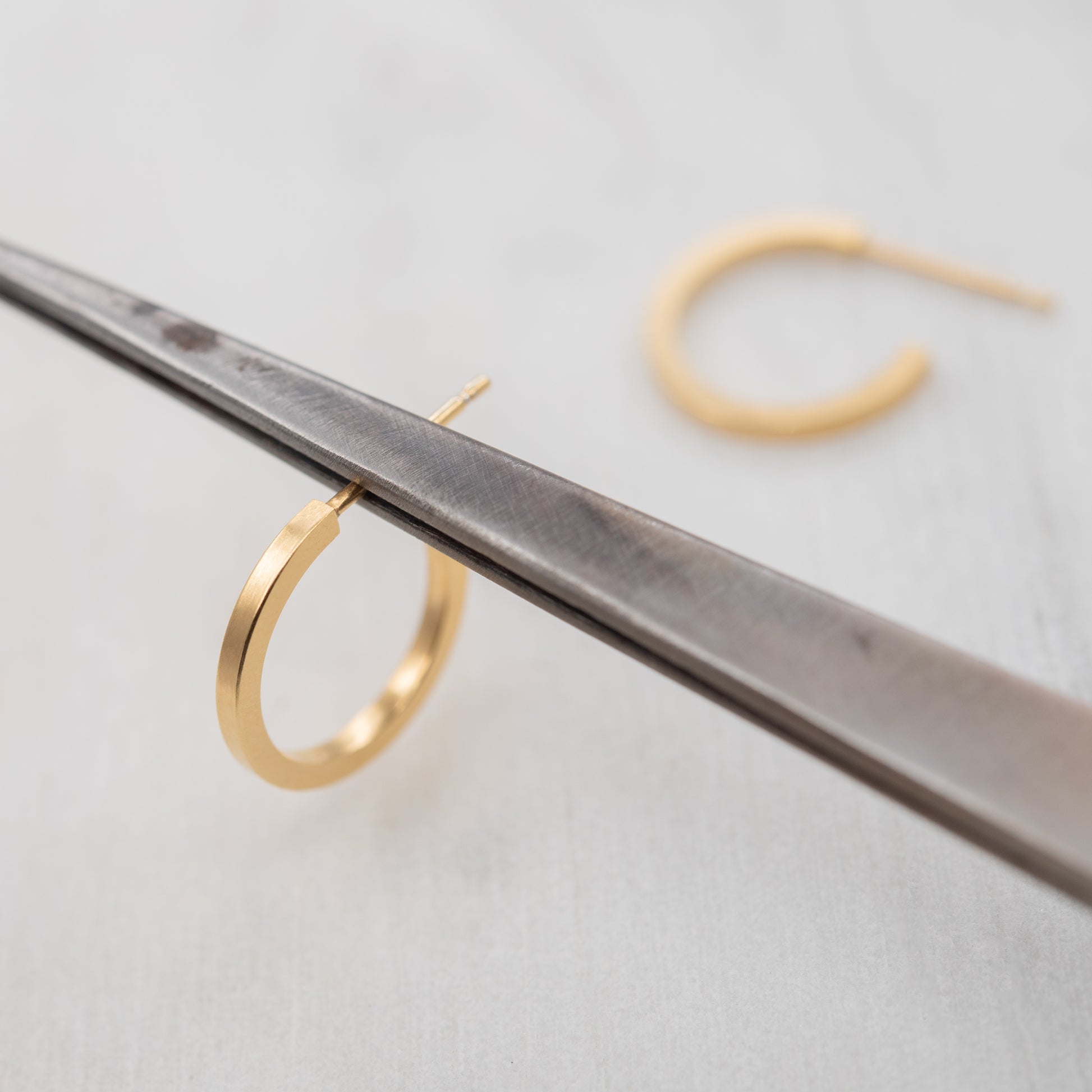 Paire de petites créoles dorées à l’or fin 24k photographiées sur un outil de bijoutier, fabrication artisanale parisienne.