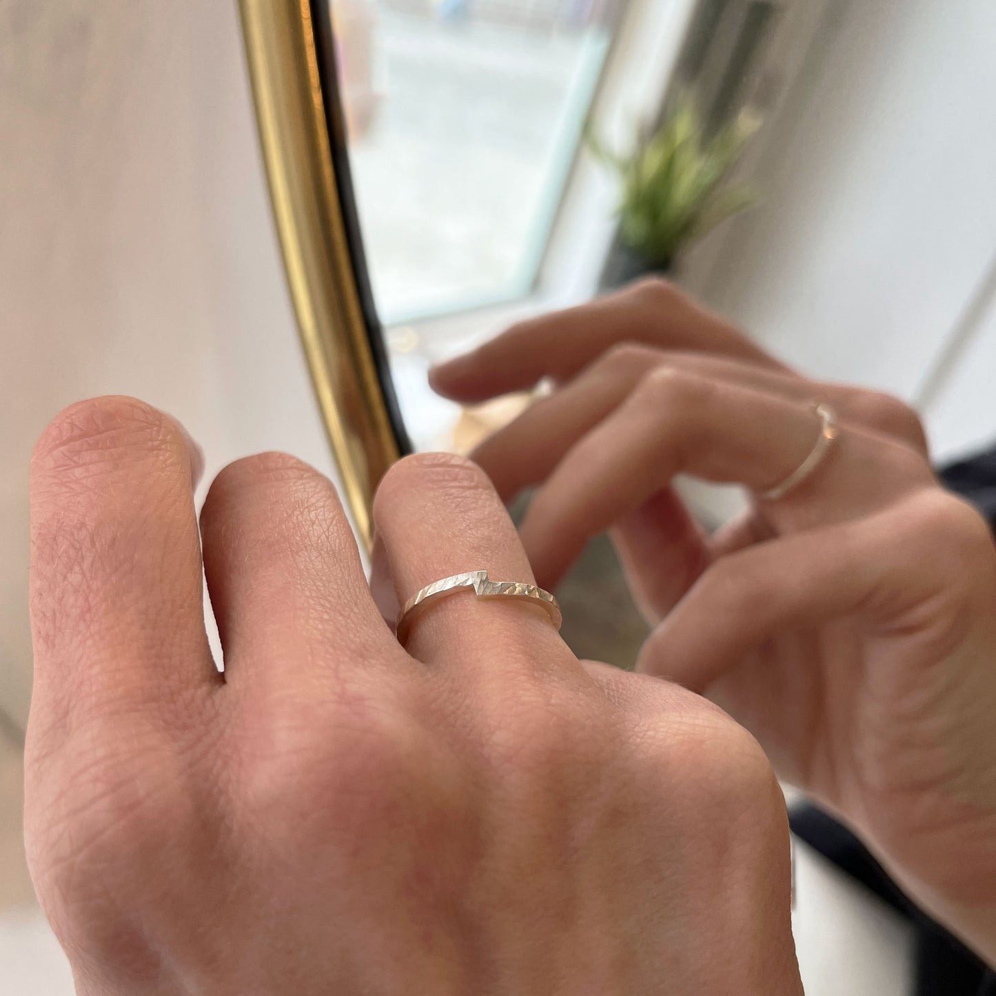Hand wearing a hammered sterling silver unity ring, with a unique design symbolizing the merging of two elements, reflected in a mirror. The ring's distinctive notch detail represents connection and harmony, making it a meaningful choice as a wedding band, engagement ring, or everyday accessory. A modern, minimalistic piece crafted by hand.