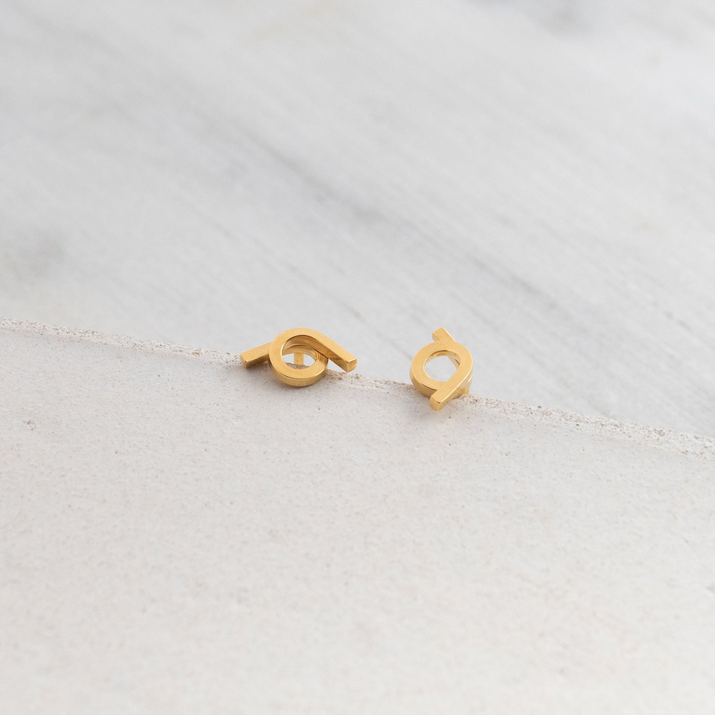 Close-up of a pair of Vermeil stud earrings showcasing a unique intertwined design. The earrings feature a shiny finish and are displayed on a textured surface, highlighting their modern and minimalist style.