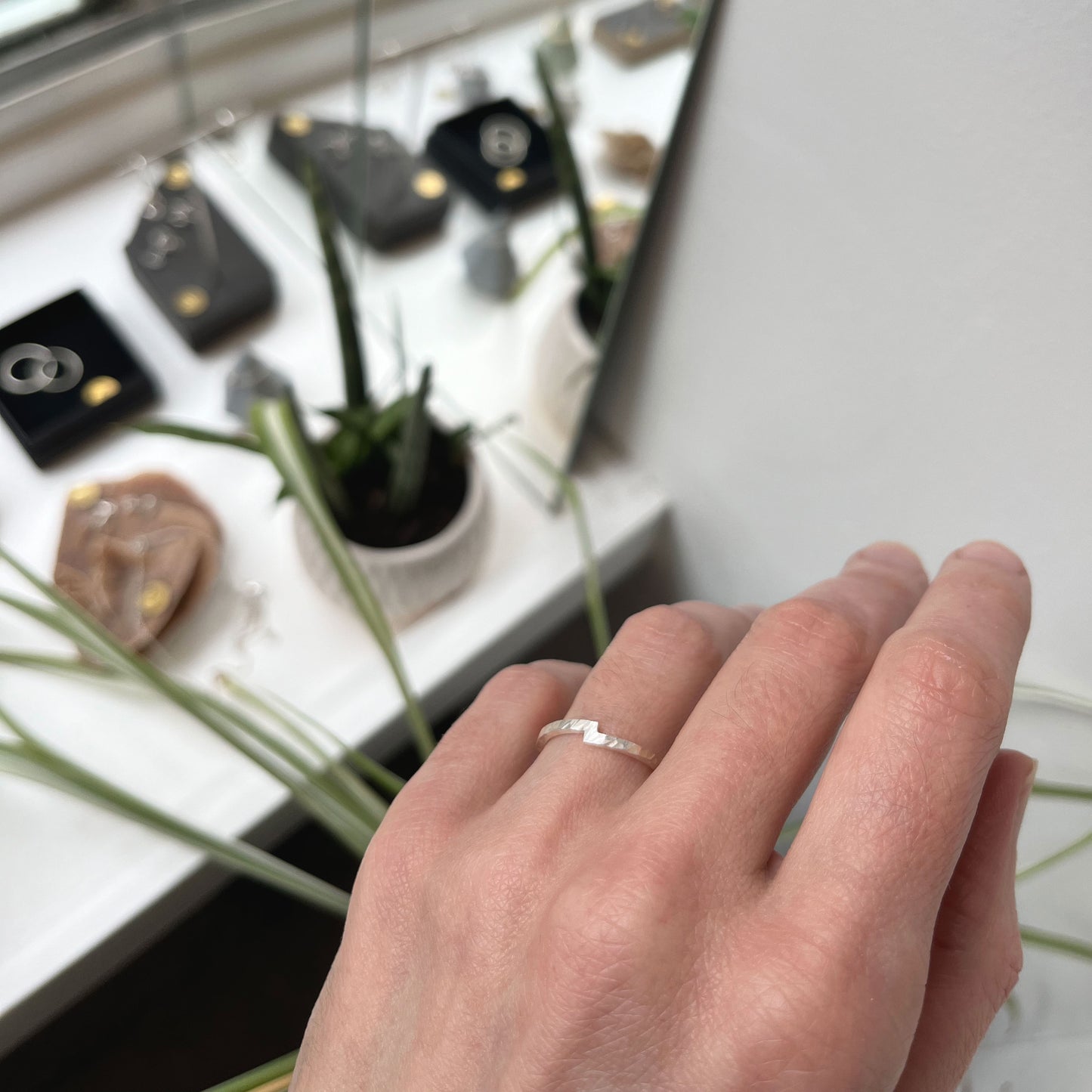 Main portant une bague en argent sterling martelée, symbolisant l'union de deux éléments avec un design unique où les extrémités se rejoignent pour représenter la connexion et l'harmonie. La bague, élégante et minimaliste, est un choix parfait pour une alliance, une bague de fiançailles ou un accessoire quotidien. En arrière-plan, un studio de bijouterie avec des pièces exposées.