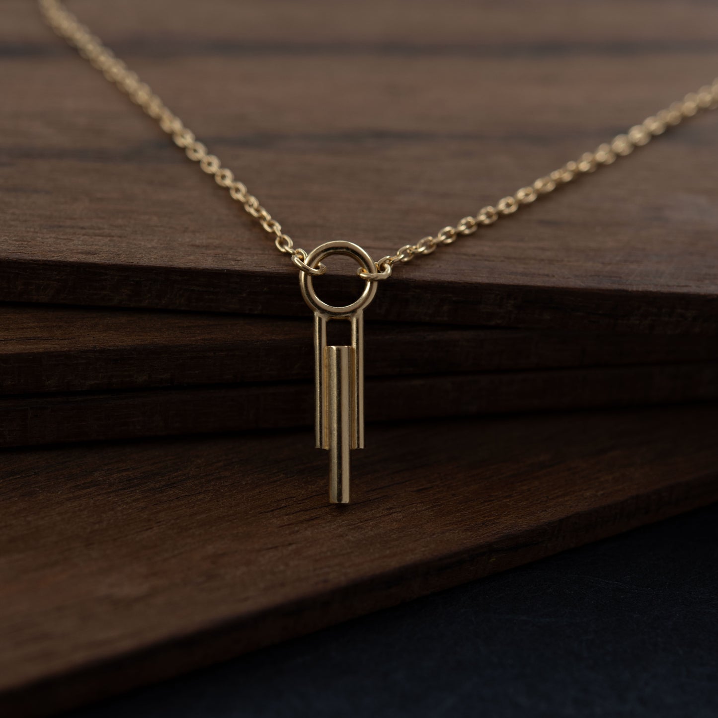 Gros plan d'un pendentif cylindrique Art Déco à trois barres avec chaîne en vermeil contre fond de bois foncé, bijou fait main à Paris