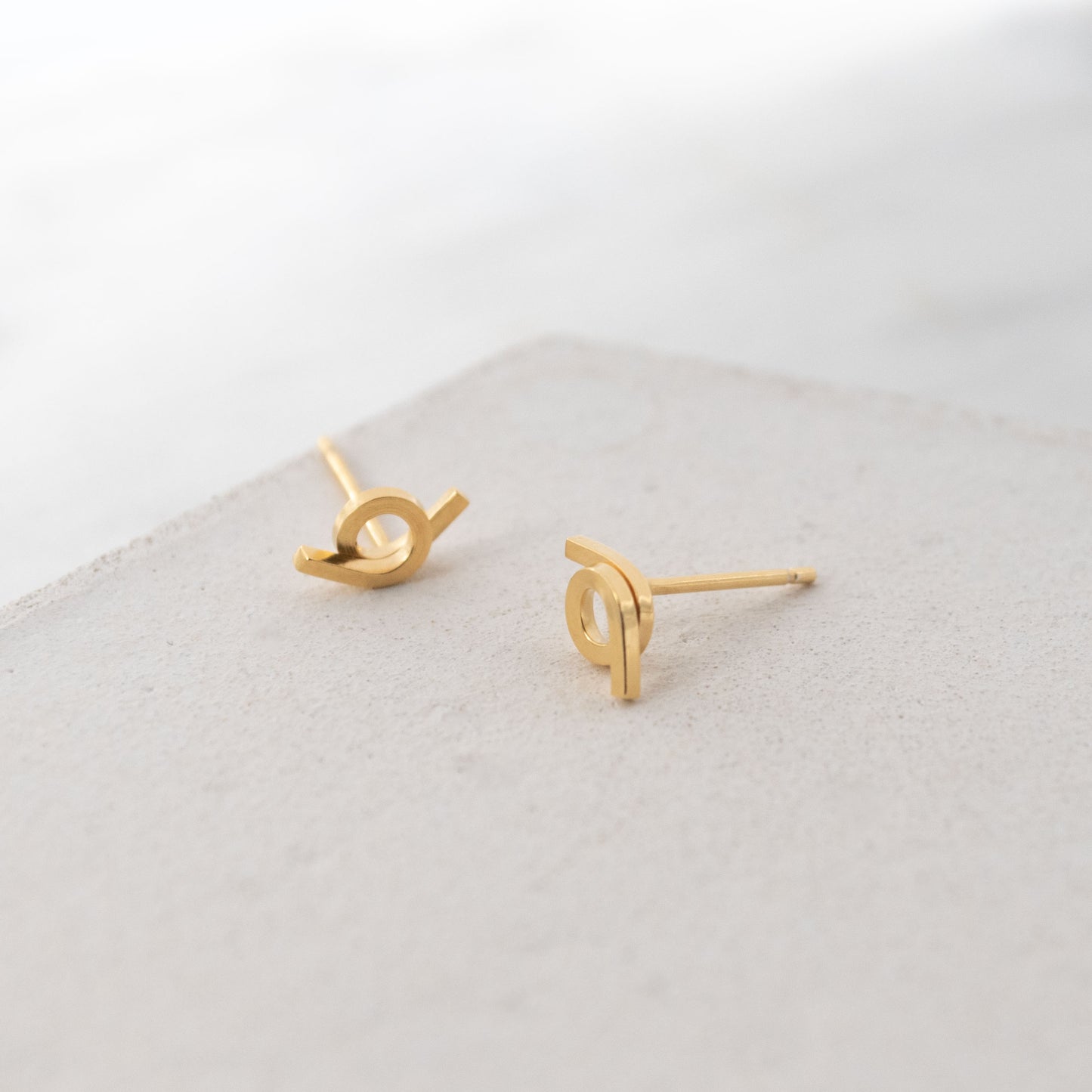 Vista cercana de un par de pendientes en vermeil con un diseño entrelazado, colocados sobre una superficie texturizada. Los pendientes muestran un acabado brillante y un estilo moderno, ideales para un look minimalista.