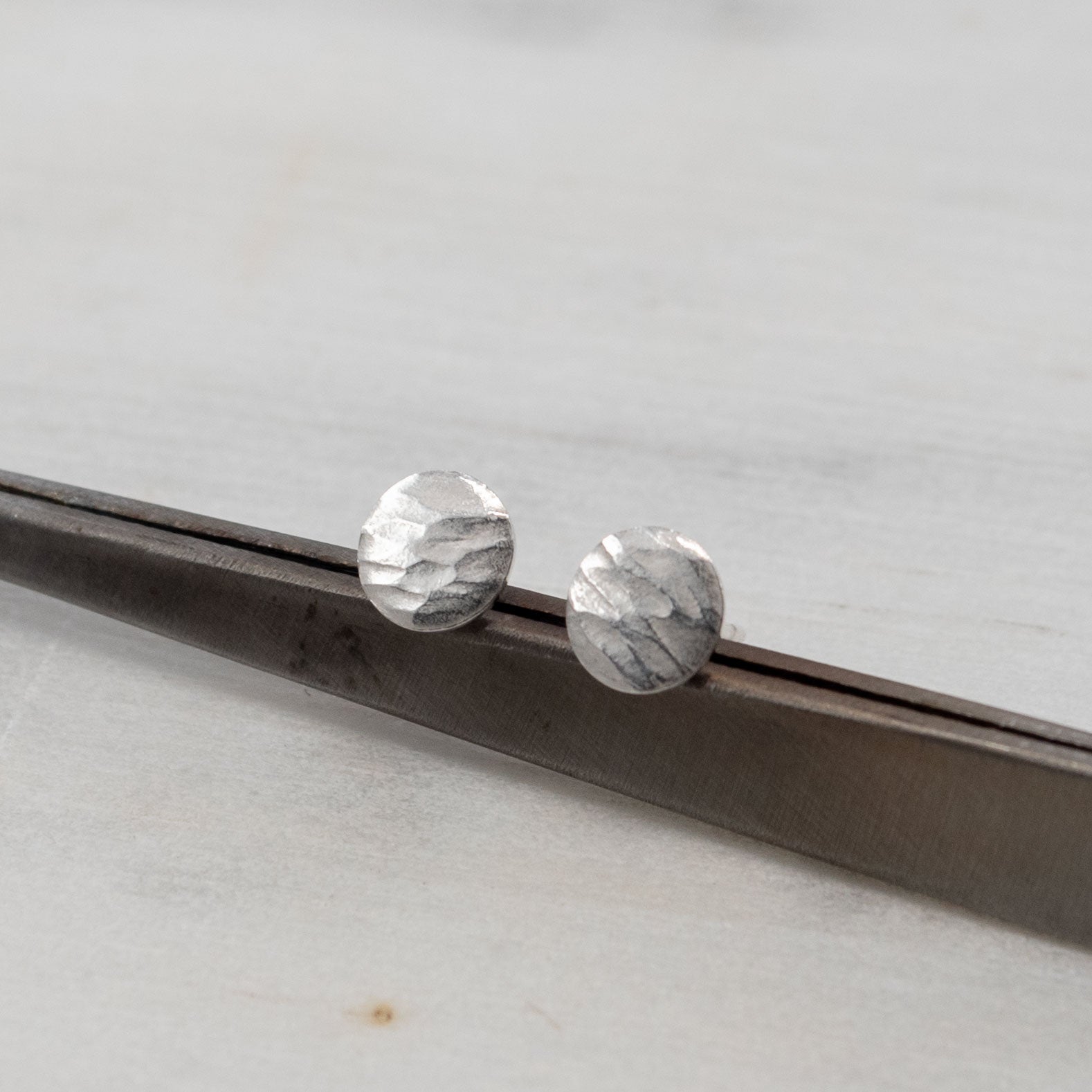Une paire de petites boucles d'oreilles en argent sterling martelé, présentée sur une surface métallique sombre. Les boucles d'oreilles ont une texture artisanale unique, avec des reflets doux qui accentuent leur finition mate et minimaliste. L'image met en valeur le design épuré et élégant de ces bijoux, parfaits pour un look discret et moderne.