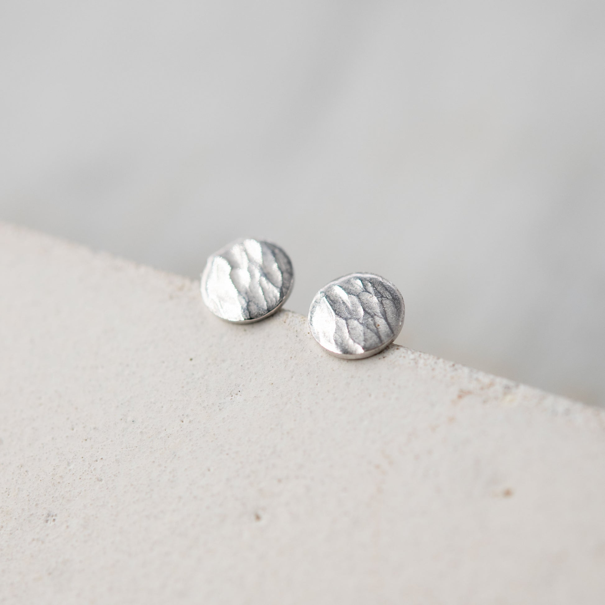 Image en gros plan de deux boucles d'oreilles en argent sterling posées sur un bord de surface claire. Les boucles d'oreilles ont une forme ronde avec une finition martelée qui capte la lumière, mettant en valeur leur texture artisanale. L'arrière-plan est flou et doux, accentuant l'élégance des bijoux.