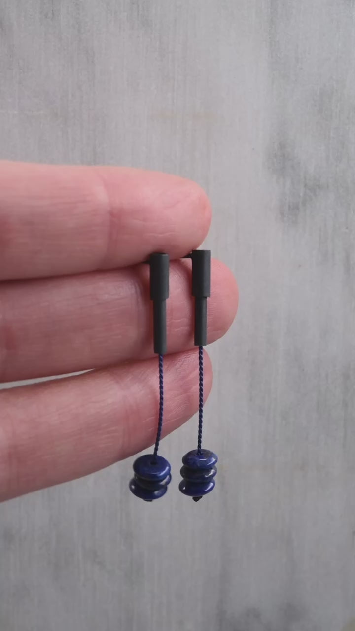 Vidéo en gros plan d'une paire de boucles d'oreilles pendantes fabriquées à la main à Paris. Les boucles sont en argent oxydé, ornées d'un fil de soie bleu vibrant et de pierres de lapis-lazuli. Ce design unique et élégant met en valeur la combinaison de matériaux raffinés pour un style à la fois artisanal et sophistiqué.