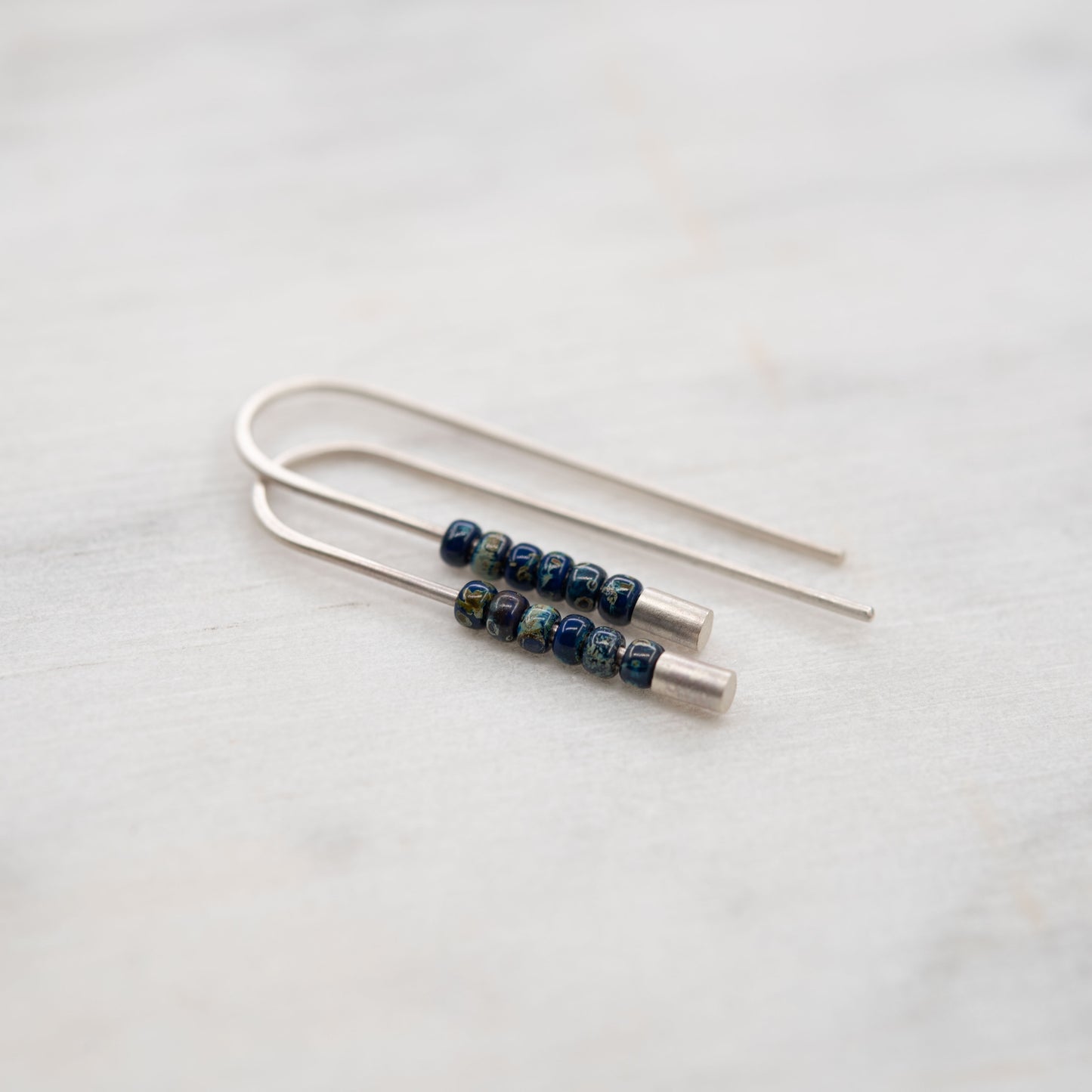 Silver threader earrings with matte finish and small blue Miyuki beads, artisan jewelry photographed on marble.