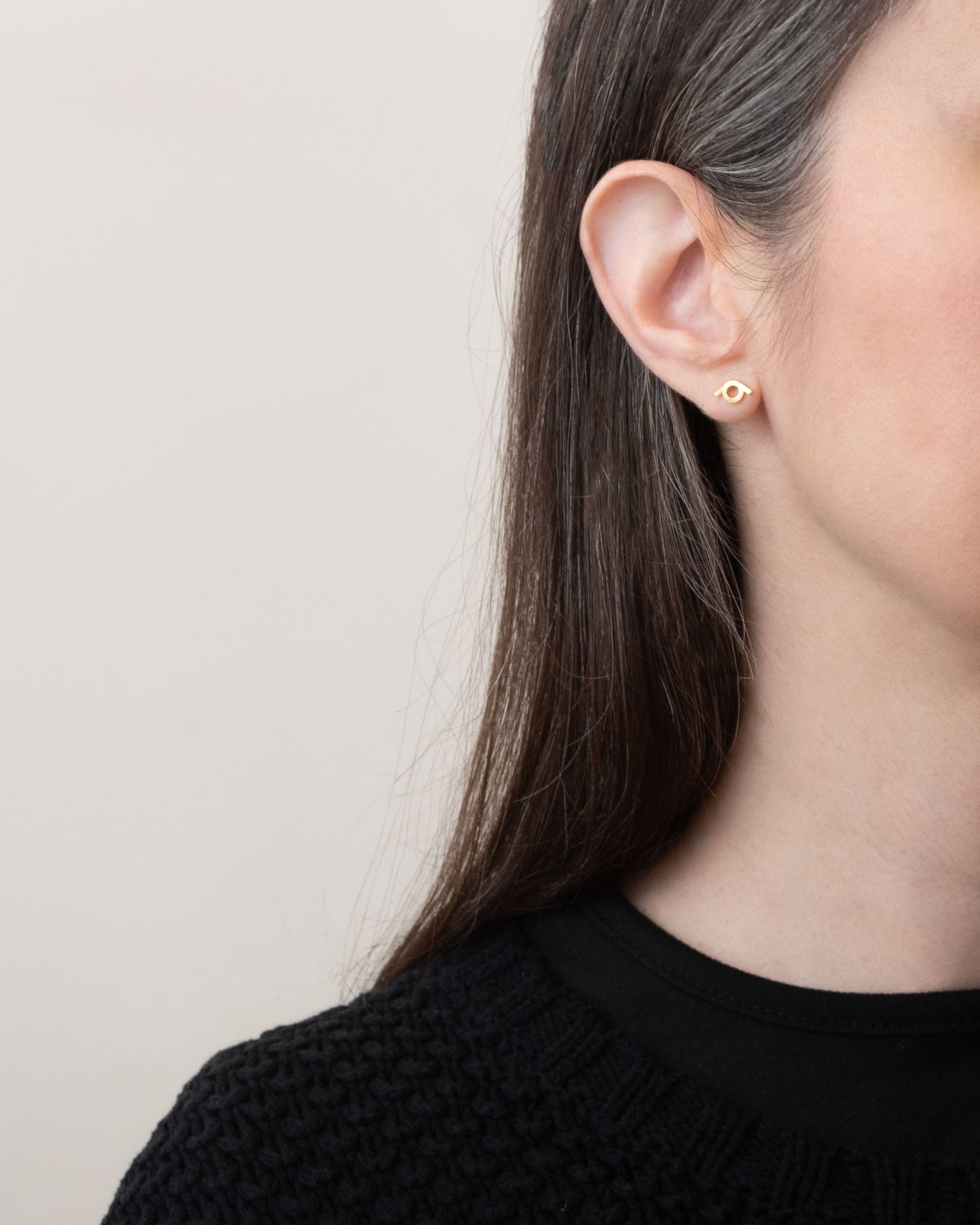 Profile view of a woman wearing a pair of Vermeil stud earrings with an intertwined design. The earrings are prominently displayed against a neutral background, and the woman has long, straight hair, wearing a black textured sweater. The image captures the earrings' modern and minimalist aesthetic.
