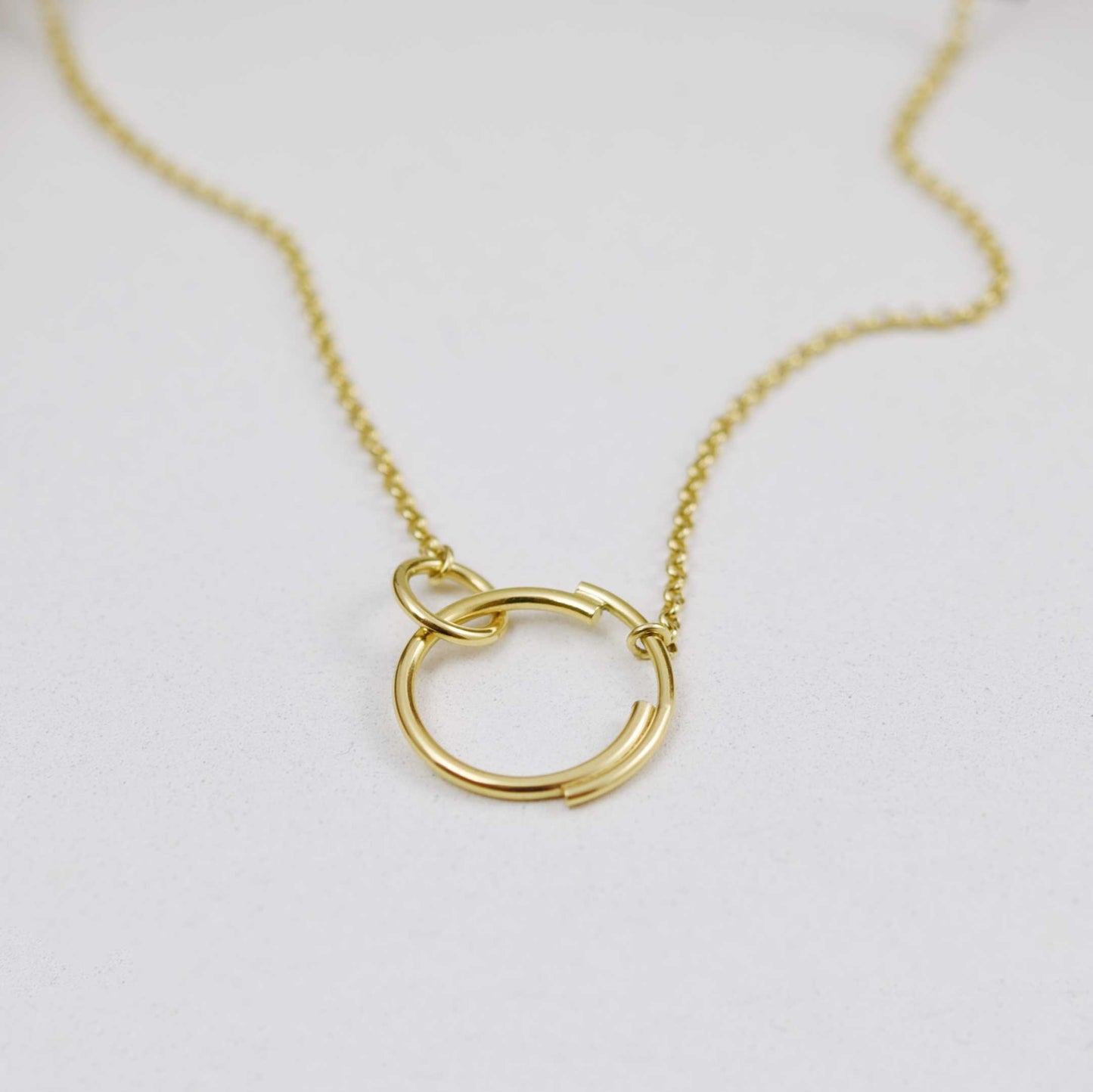 A close up of a stylish interlocking circle necklace  in gold that features a small circle clasping onto a larger circle that is made up of two separate arcs for a truly eclectic design.