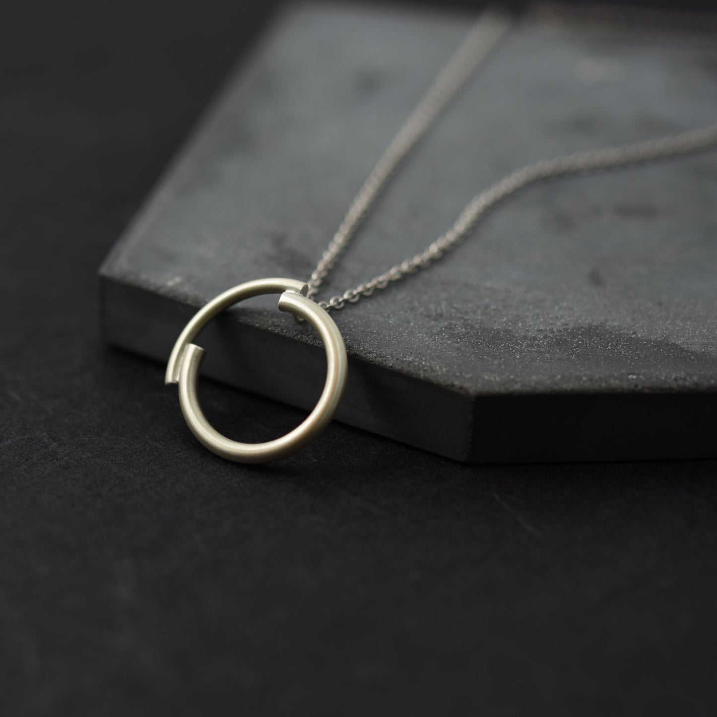 A close up of a silver circle pendant necklace of 22 millimetres diameter in a brushed matte finish in a dark blue background