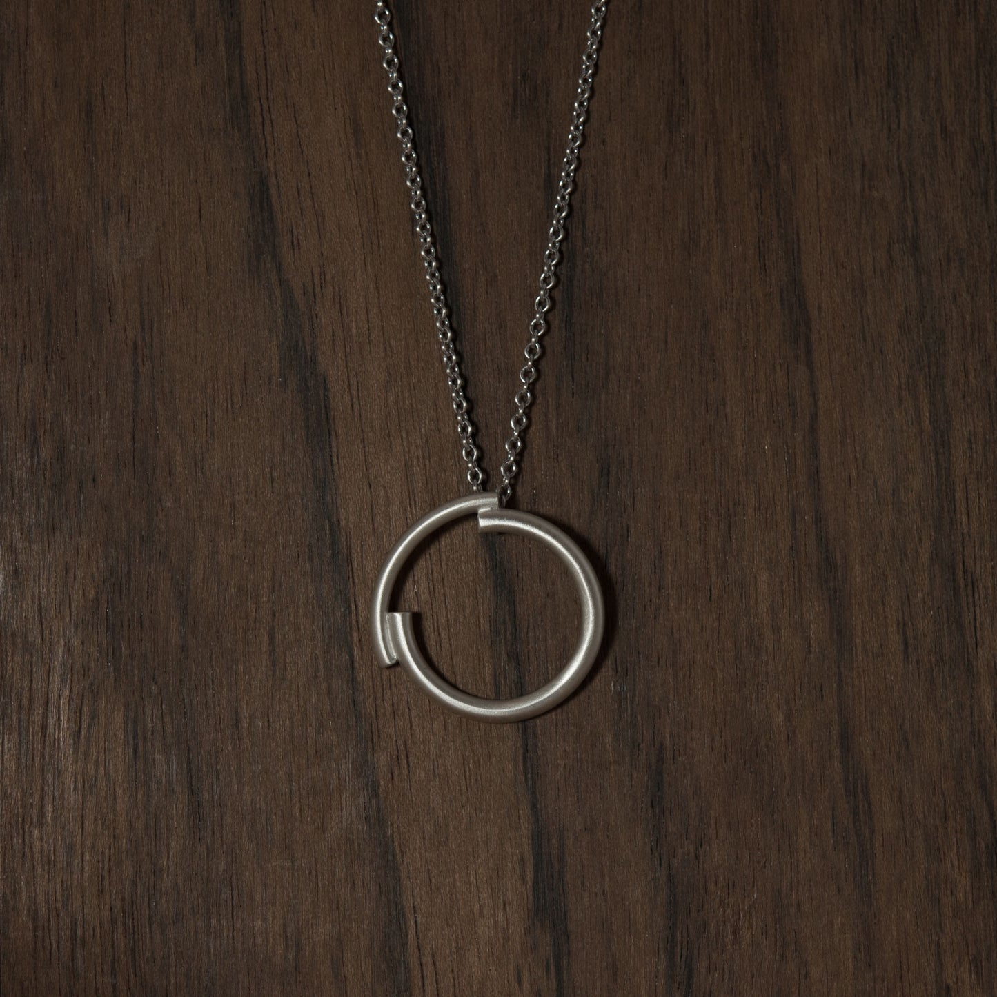 A dark wood background showing a close up of a 22 millimetres diameter circle pendant handmade in silver brush matte finished