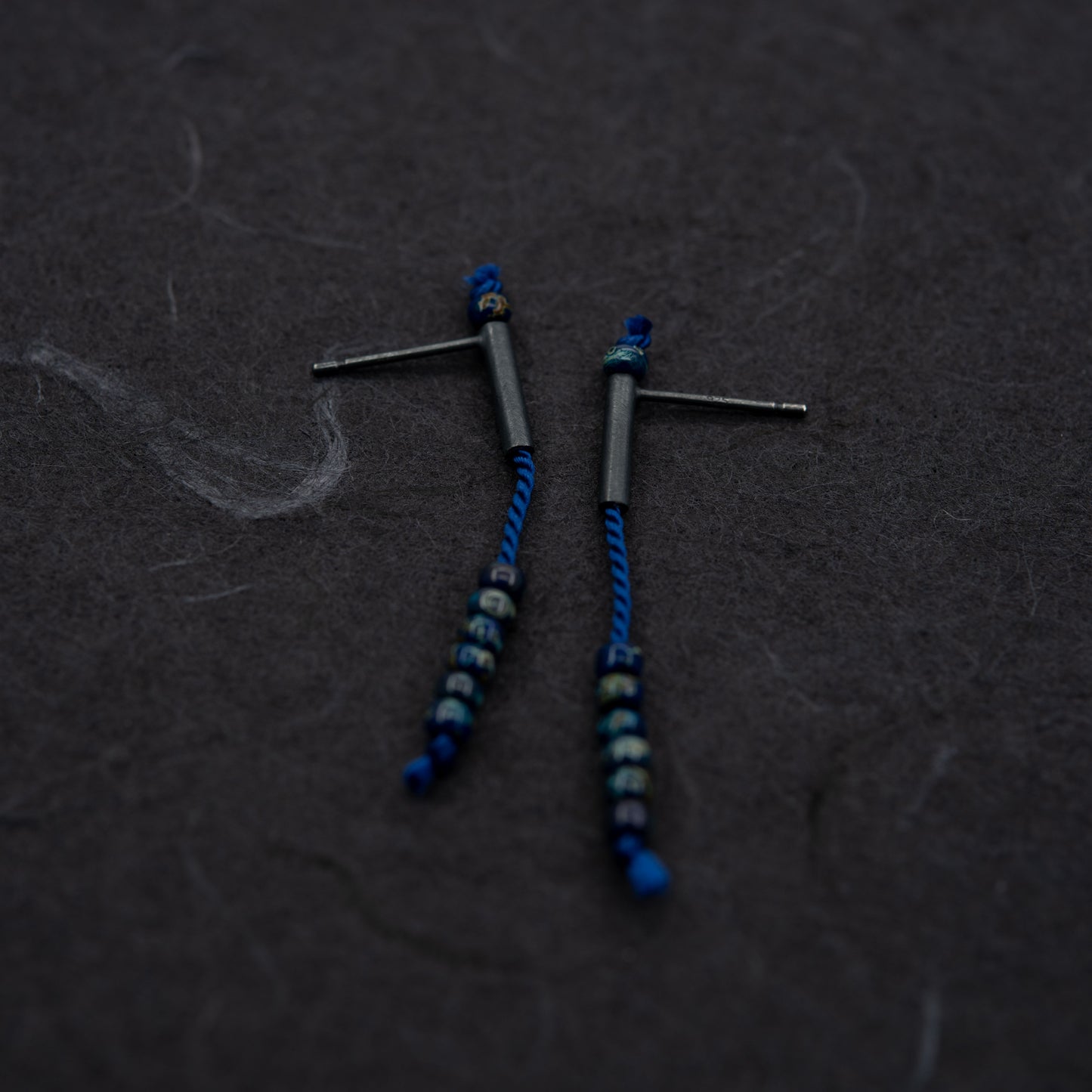 Image showing a pair of 38 mm length pendant earrings handmade in oxidized black sterling silver tube with a blue silk cord threaded through the center for hanging Picasso blue Miyuki beads. Designed and made by hand in Paris by A g J c