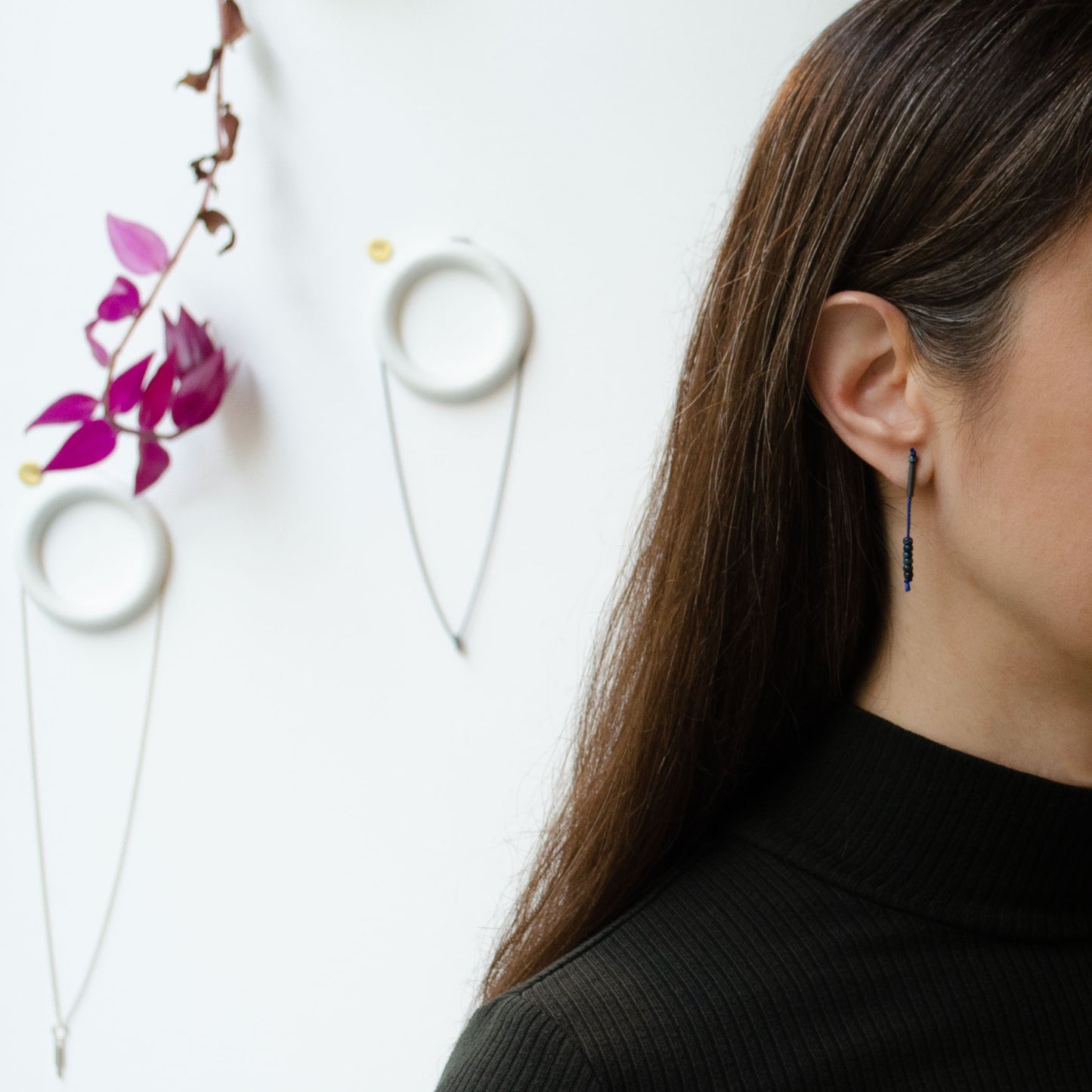 Half face of a woman wearing an earring of 38 mm length pendant earrings handmade in oxidized black sterling silver tube with a blue silk cord threaded through the center for hanging Picasso blue Miyuki beads. Made by hand in Paris by A g J c