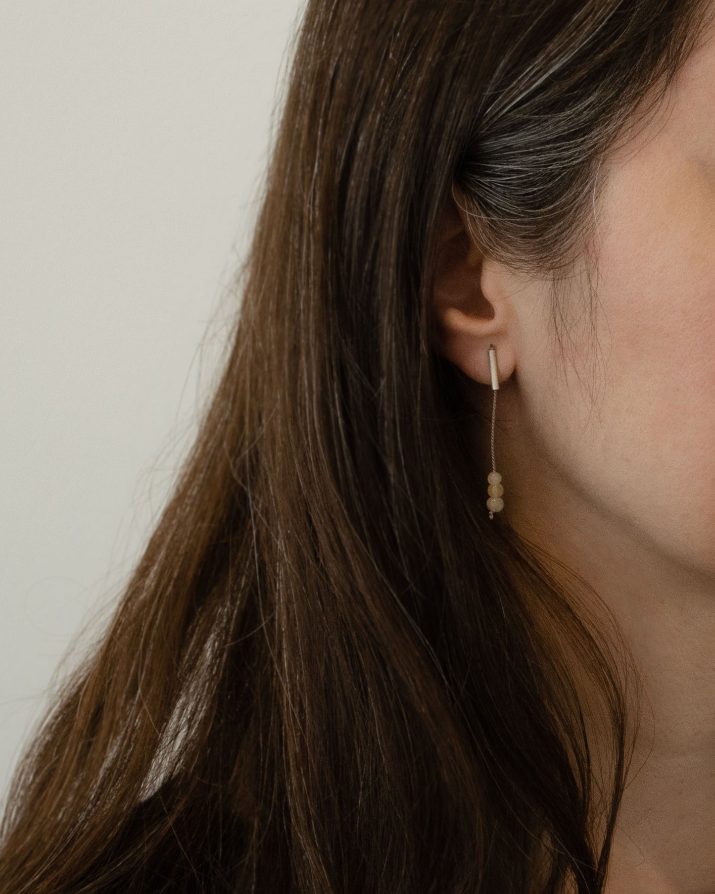 A woman wearings a pendant earring of 42 millimeters length handmade in Paris by A g J c  from  tube of matte sterling silver with a beige silk cord threaded through the center for hanging yellow quartz beads. 