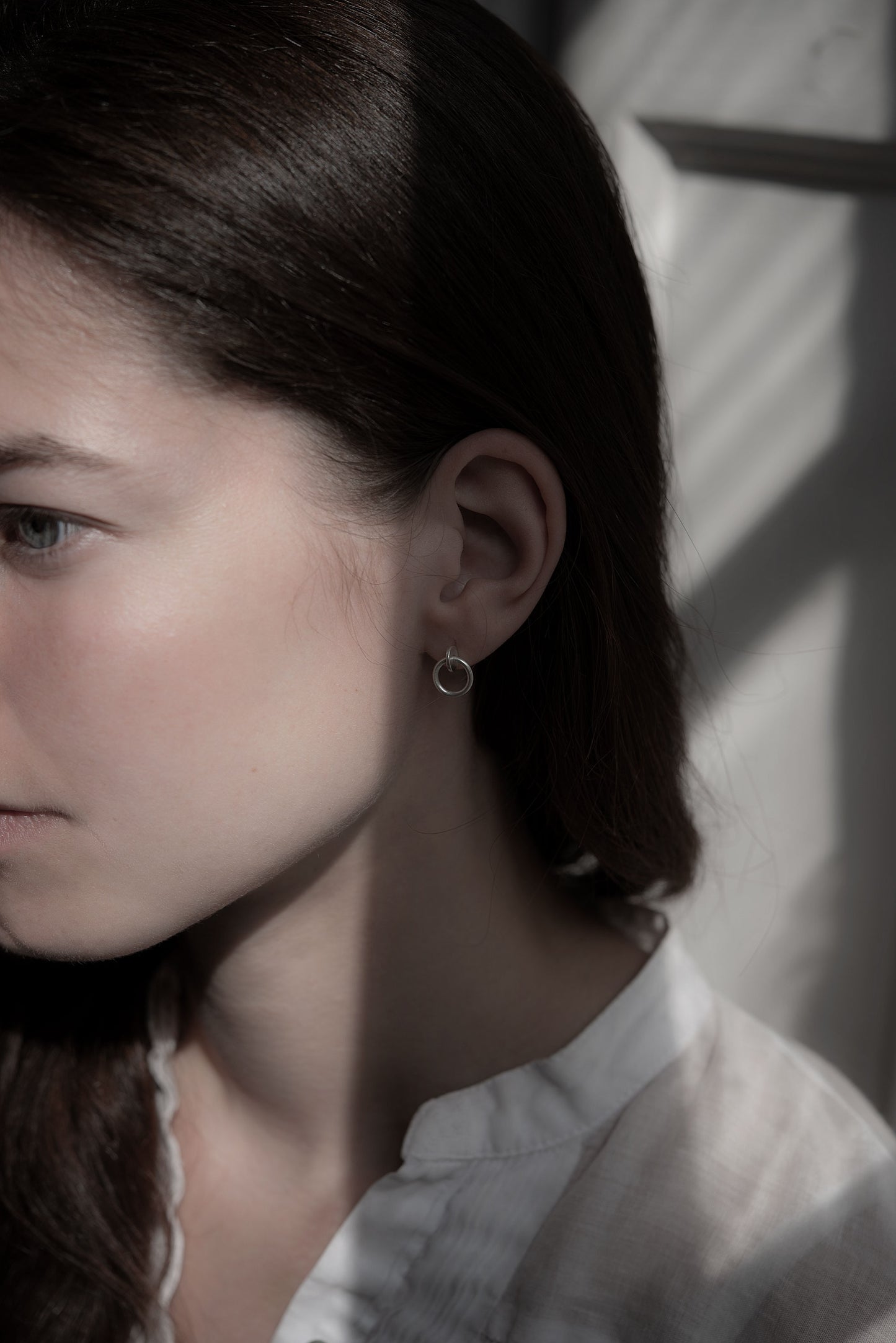 A woman's face wearing a pair of silver matte studs designed with an interlocked dot and circle The earrings measure 13 millimeters in length.