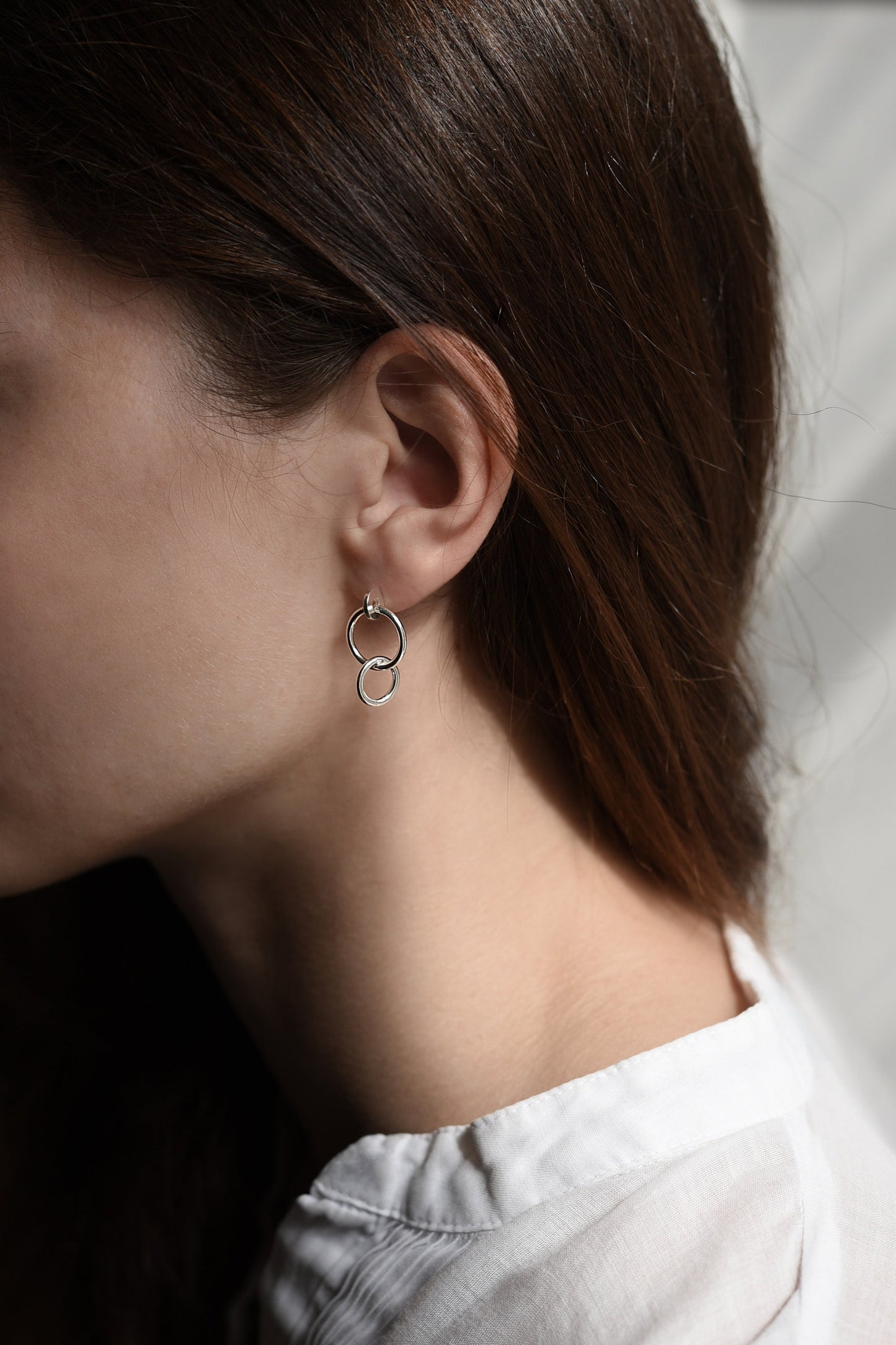 A model close to a window with a boho white shirt is wearing a pair of gold plated studs earrings designed with an interlocking dot and two hoops linked in shiny finish. Handcrafted by a g j c in Paris.