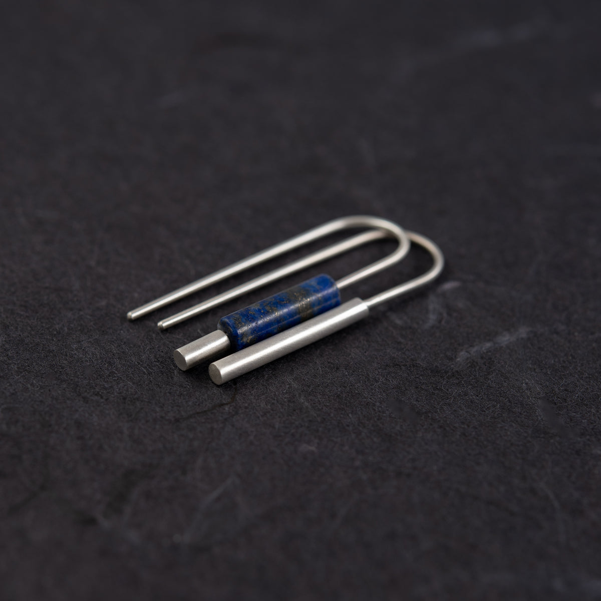 Boucles d’oreilles artisanales dépareillées en argent et lapis-lazuli vues de trois quarts, photographiée sur une surface sombre et texturée.