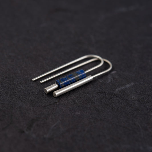 Boucles d’oreilles artisanales dépareillées en argent et lapis-lazuli vues de trois quarts, photographiée sur une surface sombre et texturée.