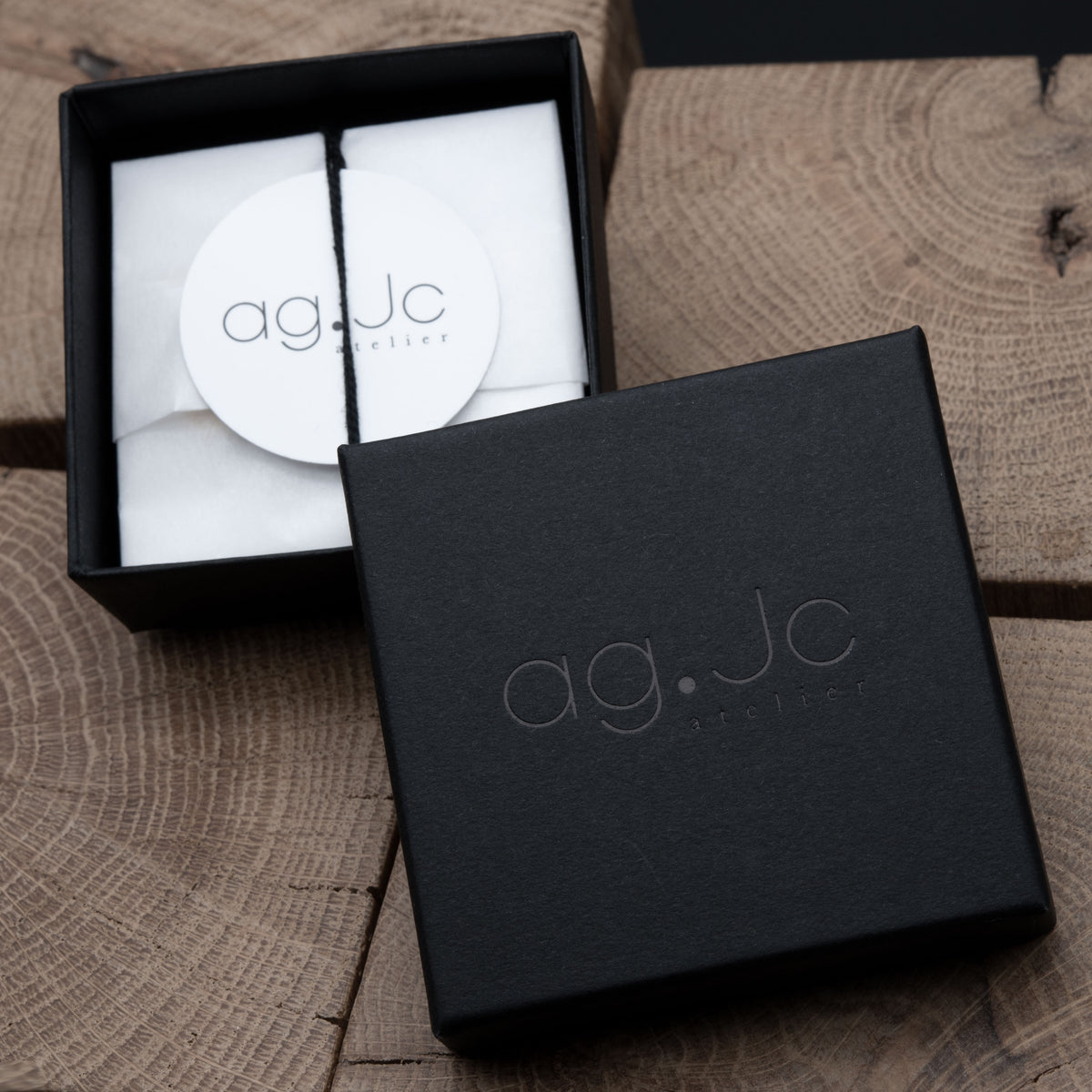 A black jewelry gift box shown open with white tissue paper and a round white tag reading 'ag.Jc jewelry', alongside the matching closed lid on a rustic wood surface.