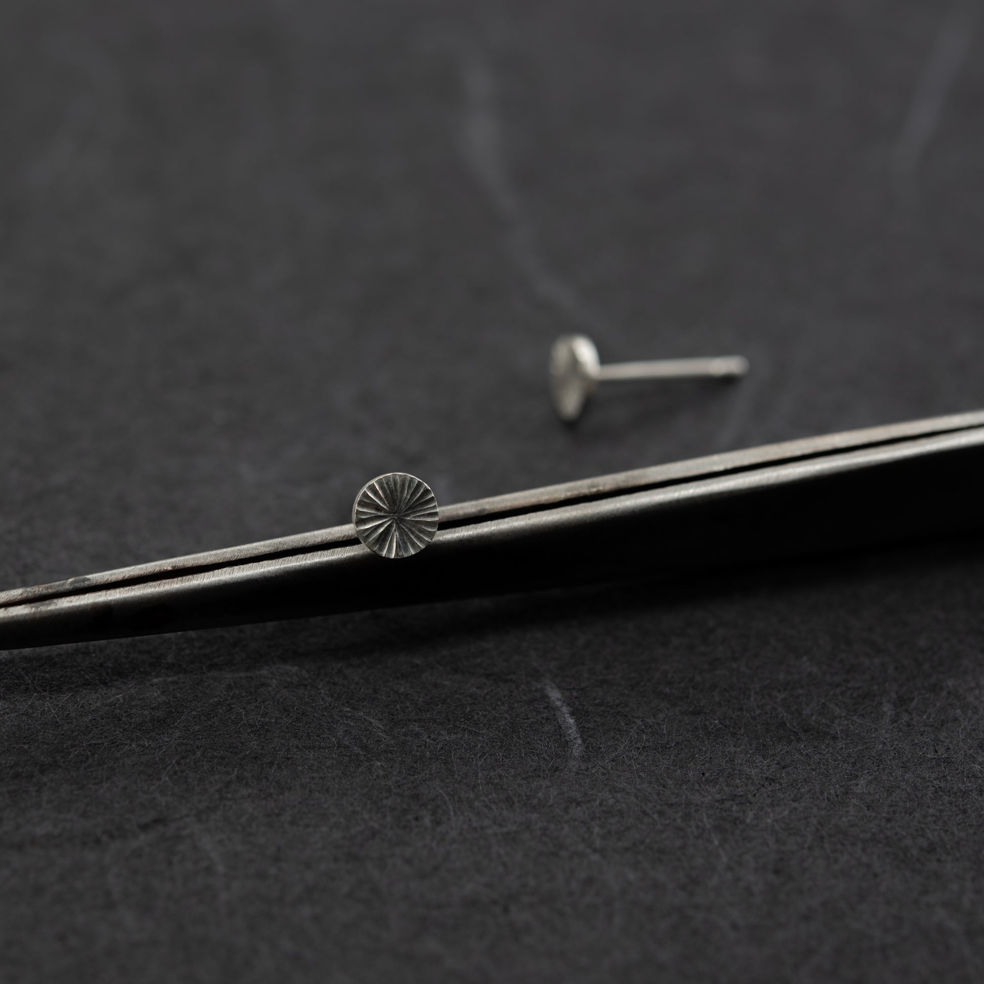 Close-up of hand-engraved sterling silver stud earrings with light-reflecting pattern on a dark textured background