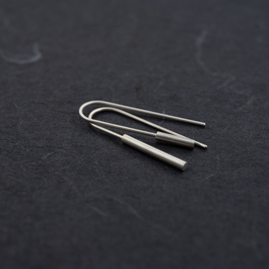 Artistic angled view of mismatched matte silver cylinder threader earrings, photographed on a black background – Paris jewelry in a clean, handmade style.
