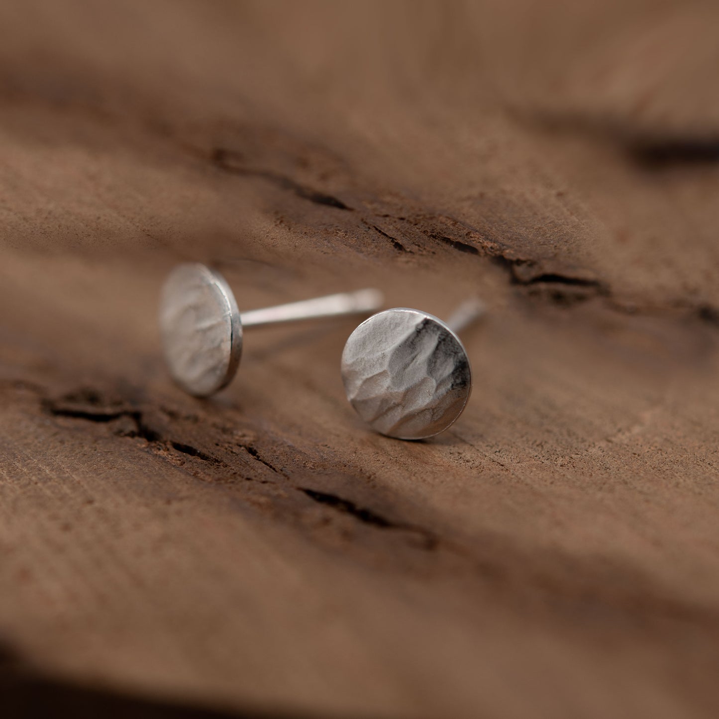 Close-up image of a pair of sterling silver stud earrings resting on a textured wooden surface. The earrings feature a round design with a hammered finish that reflects light, emphasizing their artisanal quality. The soft, natural tones of the wood provide a warm backdrop that highlights the elegance of the silver.