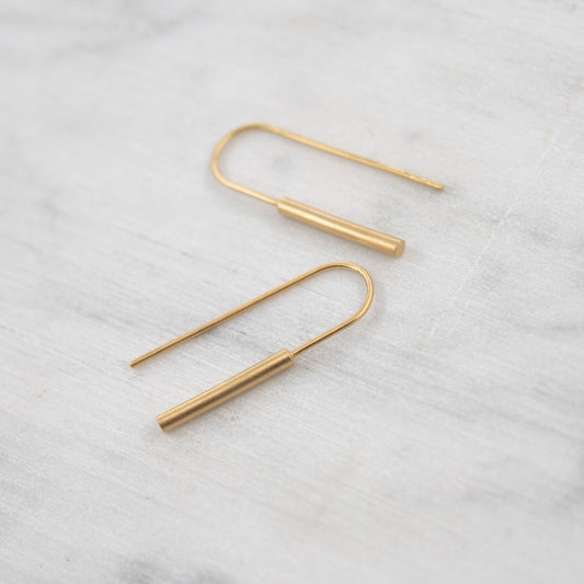 Paire de boucles d’oreilles transversantes en vermeil posées sur fond en marbre clair – design minimaliste et cylindrique en argent plaqué or 24k, finition mate