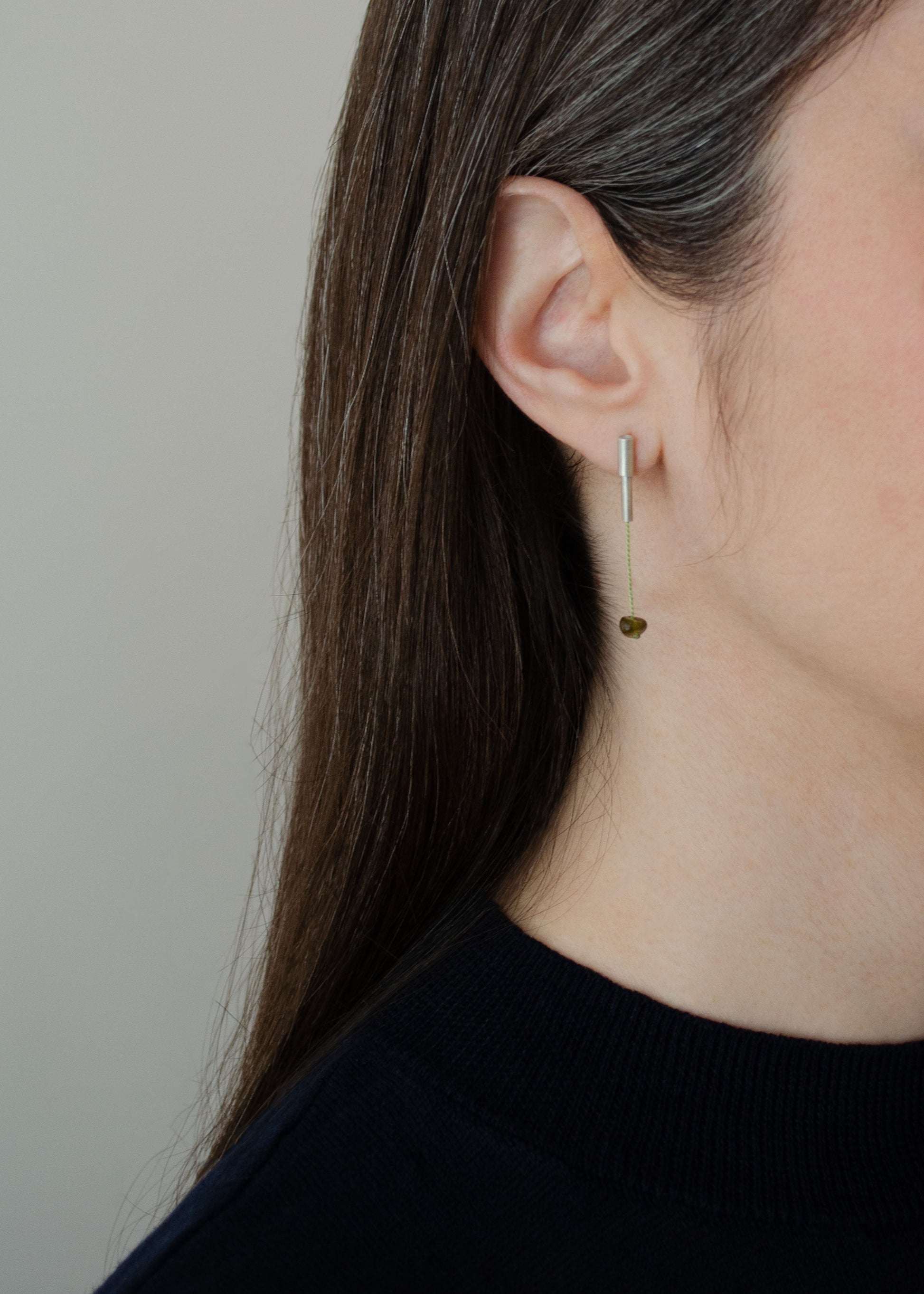 A half face of a woman wearing a pair of 37 millimeters length pendant earrings handmade in in Paris by A g J c  from  tube of matte sterling silver with a light green silk cord threaded through the center for hanging a green garnet stone.