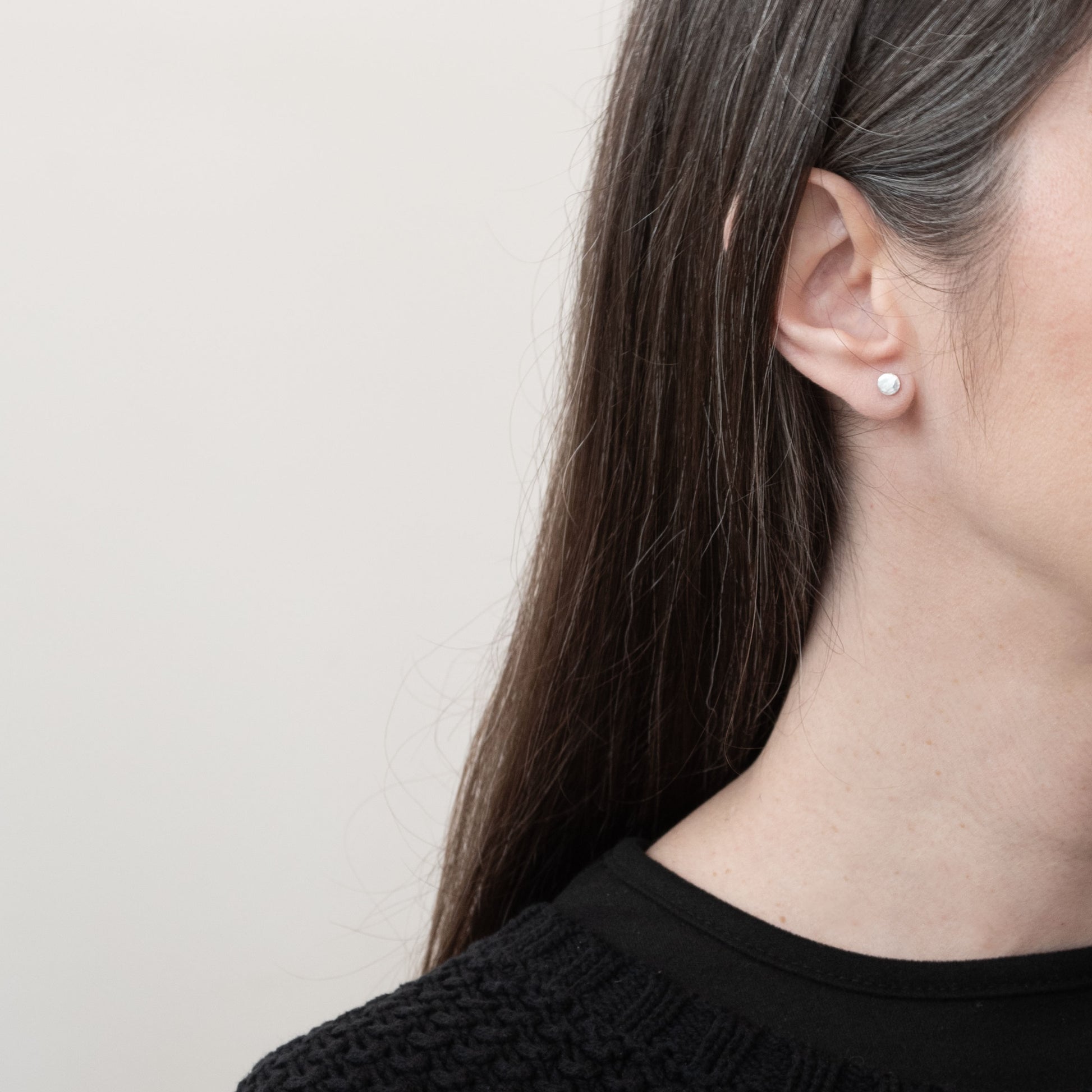 Close-up of a woman's ear wearing a single small round sterling silver stud earring with a matte, slightly textured finish. The earring has a minimalist and understated design that complements the simplicity of her long, dark hair and the black sweater she's wearing. The background is a soft off-white, adding a clean, modern look to the image.