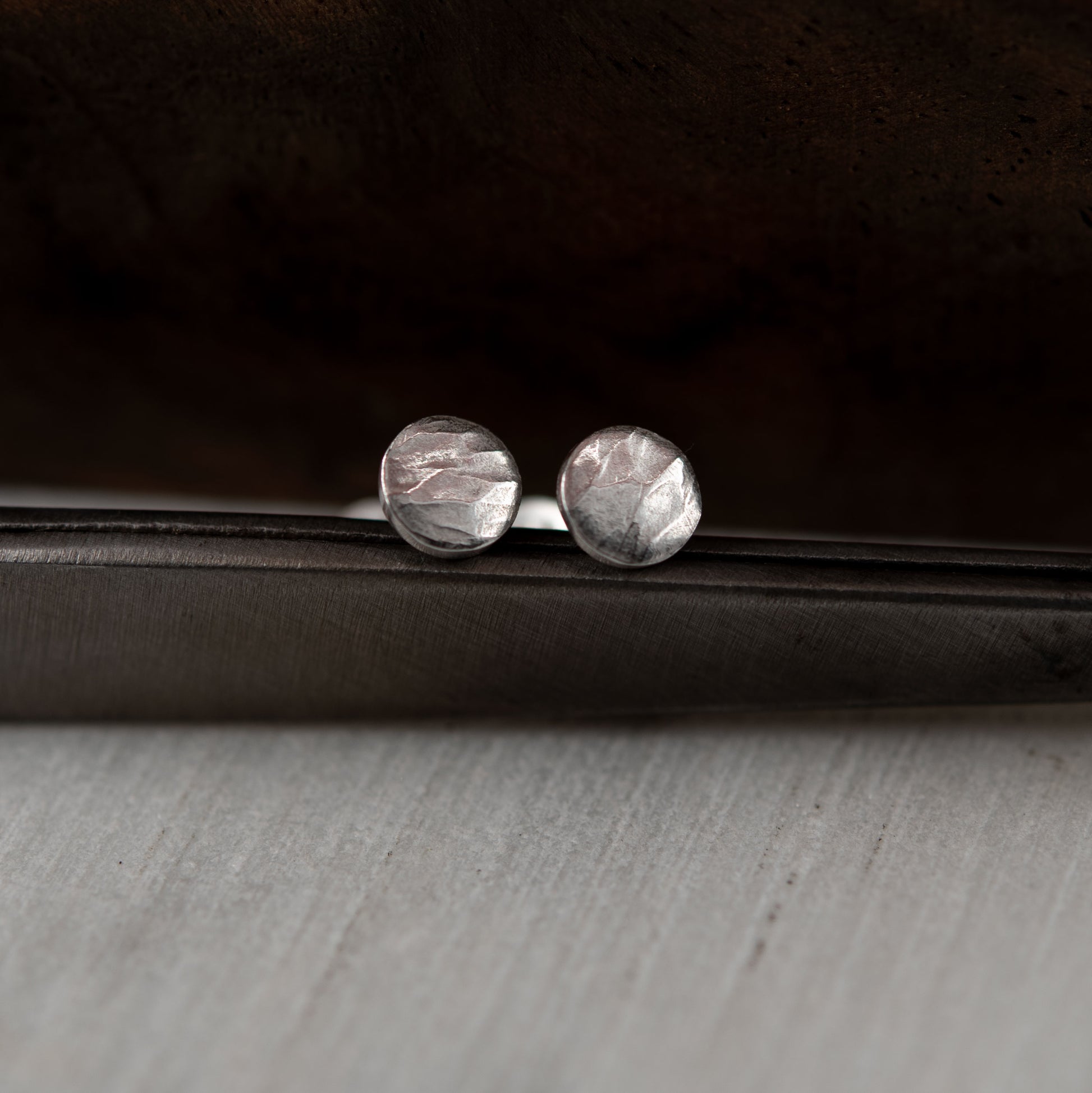 A pair of handcrafted round stud earrings made from sterling silver with a hammered texture, giving them a unique, rustic appearance. The earrings have a soft, matte finish, with subtle reflections highlighting the intricate surface details. They are displayed on a dark background, enhancing their artisanal, minimalist design.