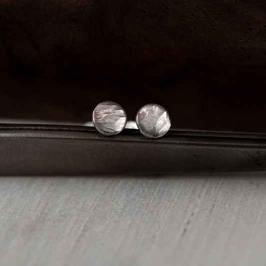 A pair of handcrafted round stud earrings made from sterling silver with a hammered texture, giving them a unique, rustic appearance. The earrings have a soft, matte finish, with subtle reflections highlighting the intricate surface details. They are displayed on a dark background, enhancing their artisanal, minimalist design.