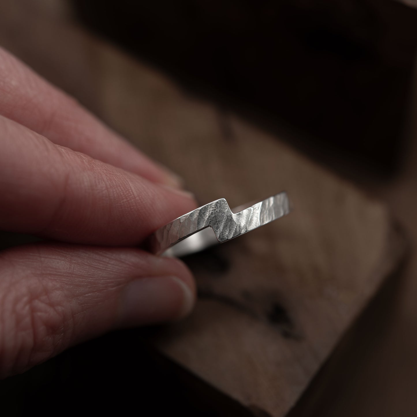 Primer plano de una mano sosteniendo el anillo a cintas cruzadas en plata esterlina. La imagen destaca el diseño único y texturizado del anillo, que se muestra sobre una superficie de madera.