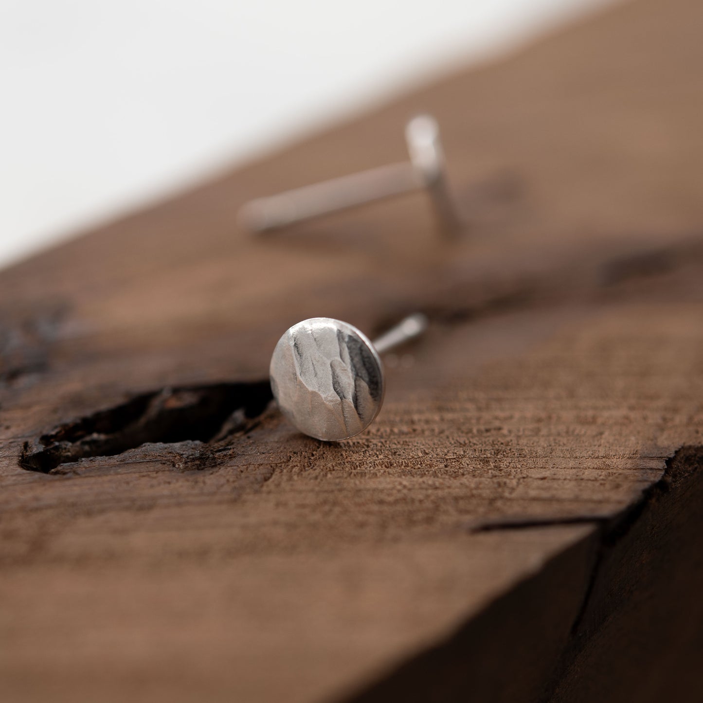 Imagen en primer plano de una pareja de pendientes de aro en plata de ley reposando sobre una superficie de madera texturizada. Los pendientes tienen un diseño redondo con una superficie martillada que refleja la luz, resaltando su calidad artesanal. El fondo suave y natural de la madera proporciona un contraste cálido que acentúa la elegancia de la plata.