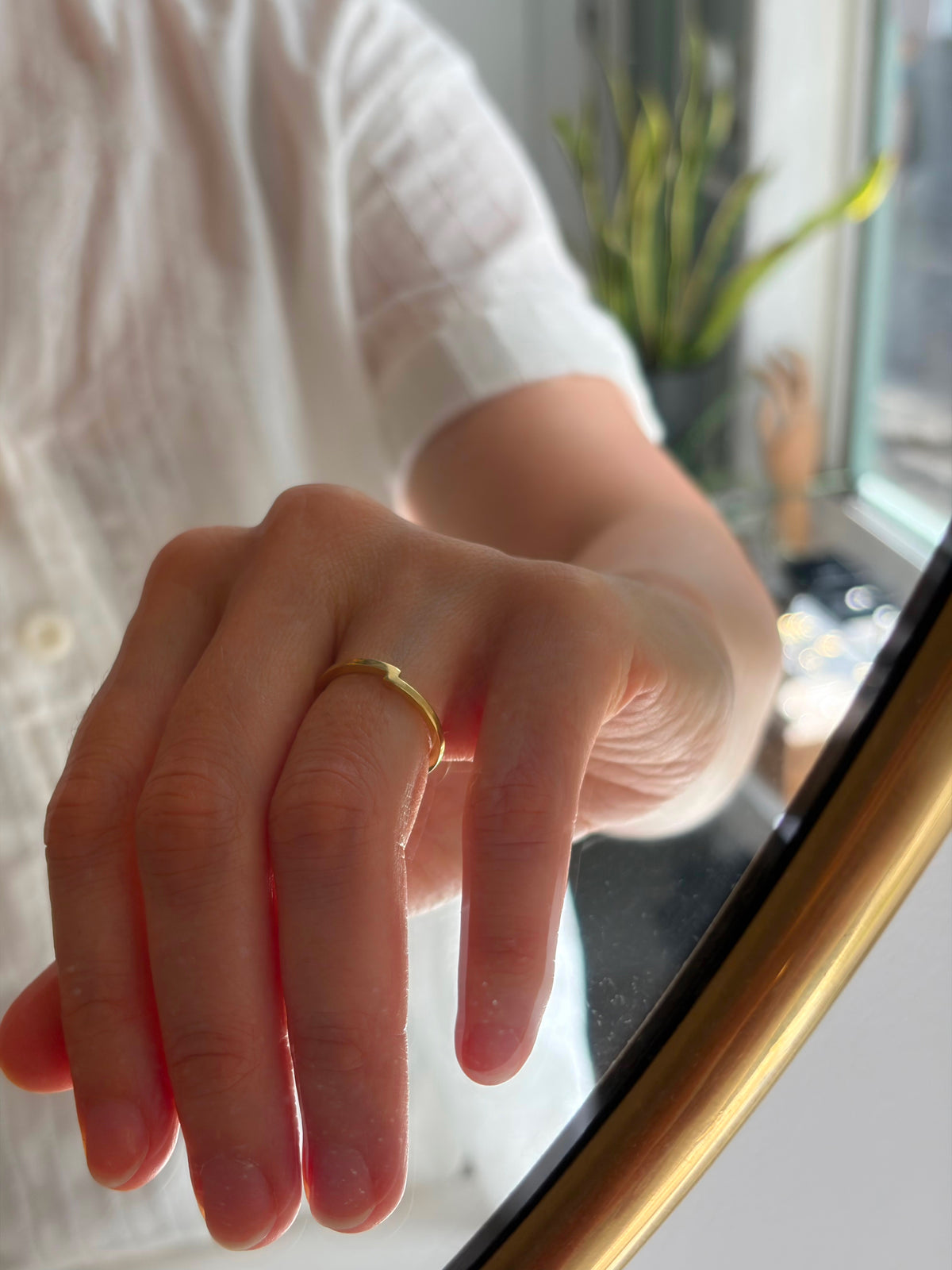 A close-up of a person's hand wearing a simple gold band ring, resting near a gold-framed mirror in a bright indoor setting.