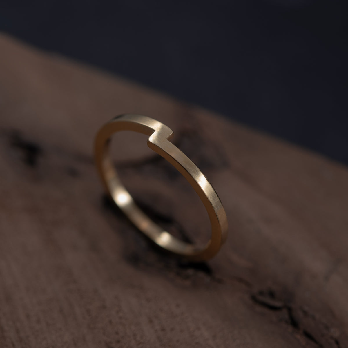 Une bague minimaliste en or avec une petite encoche sur la partie supérieure de l’anneau, posée sur une surface en bois sombre texturé.