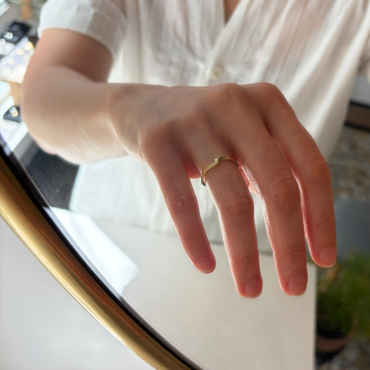Gros plan d’une main portant une fine bague symbolique en or poli, posée près d’une surface en verre réfléchissante avec un cadre doré.