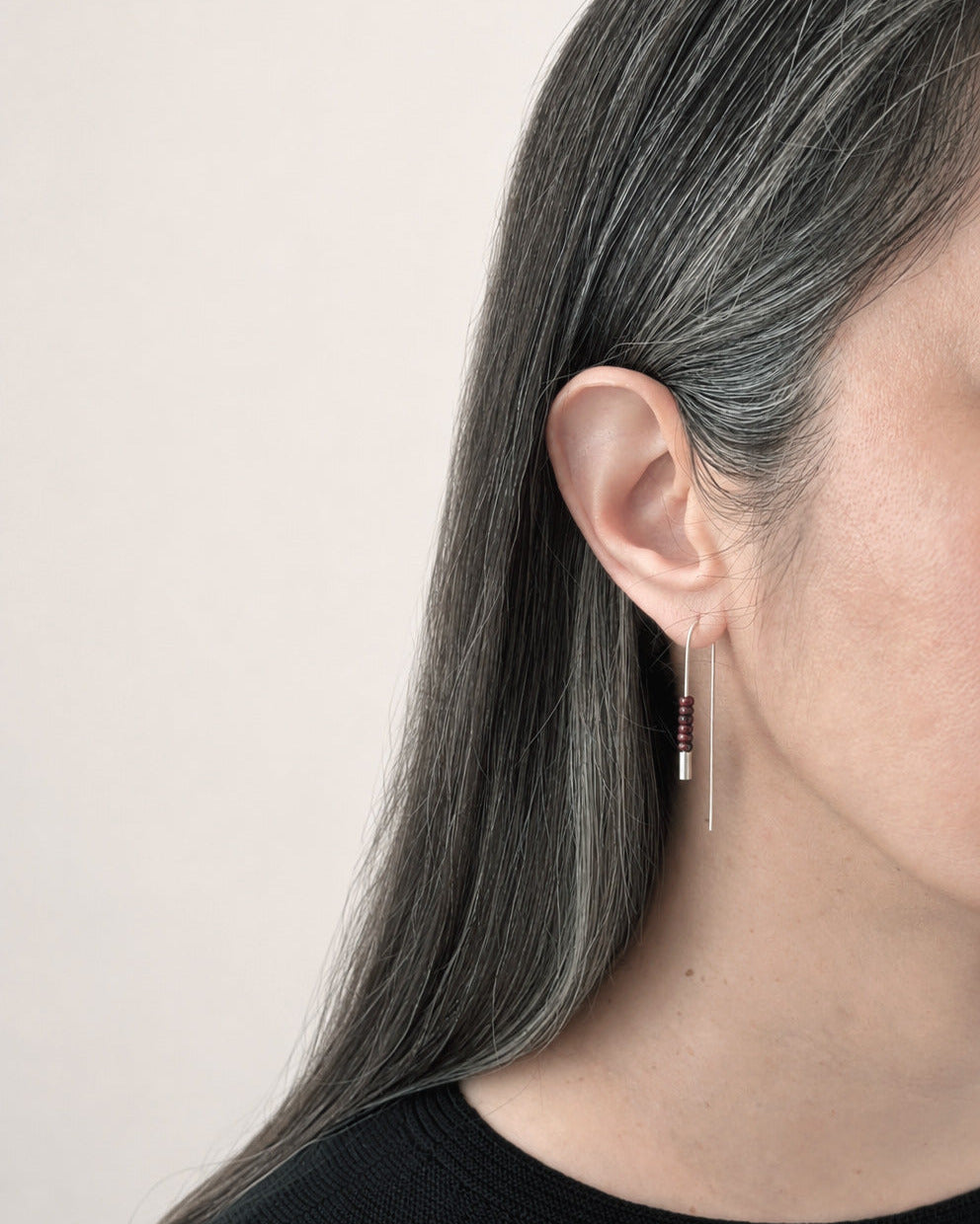 Un gros plan de profil d’une femme aux longs cheveux gris foncé, portant une délicate boucle d’oreille pendante en argent ornée de petites perles rouges, sur un fond uni clair.