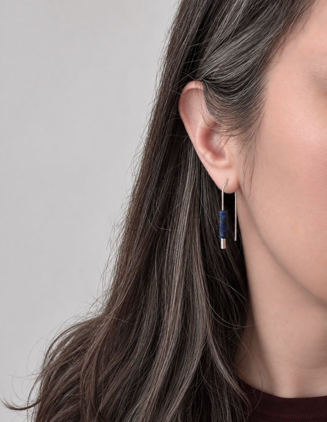 Gros plan sur l’oreille d’une femme portant une longue boucle d’oreille pendante minimaliste, composée d’une perle cylindrique bleu foncé et d’un élément pendant en argent, sur un fond gris clair.