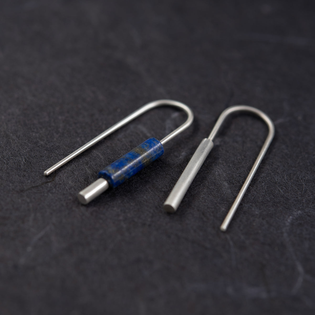 Boucles d’oreilles asymétriques en argent mat et lapis-lazuli naturel sur fond sombre.