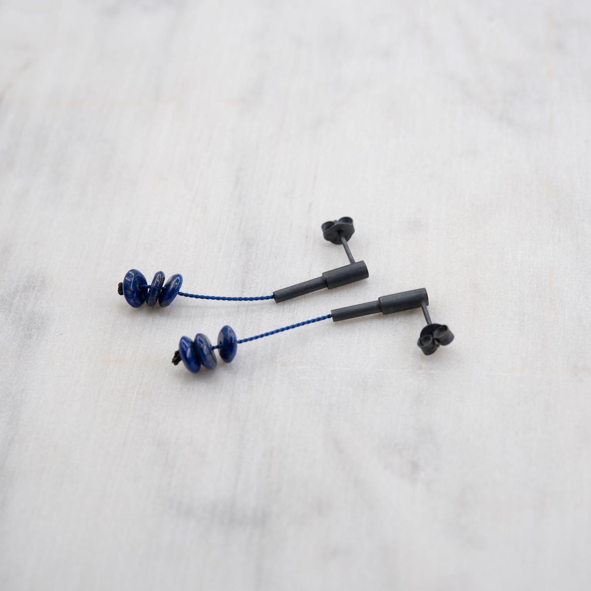 Vue rapprochée des boucles d'oreilles en lapis lazuli avec un design en argent oxydé et cordon en soie bleu, posées sur une surface en marbre. Les perles bleues sont empilées, mettant en valeur leur éclat et l'élégance moderne de l'accessoire.