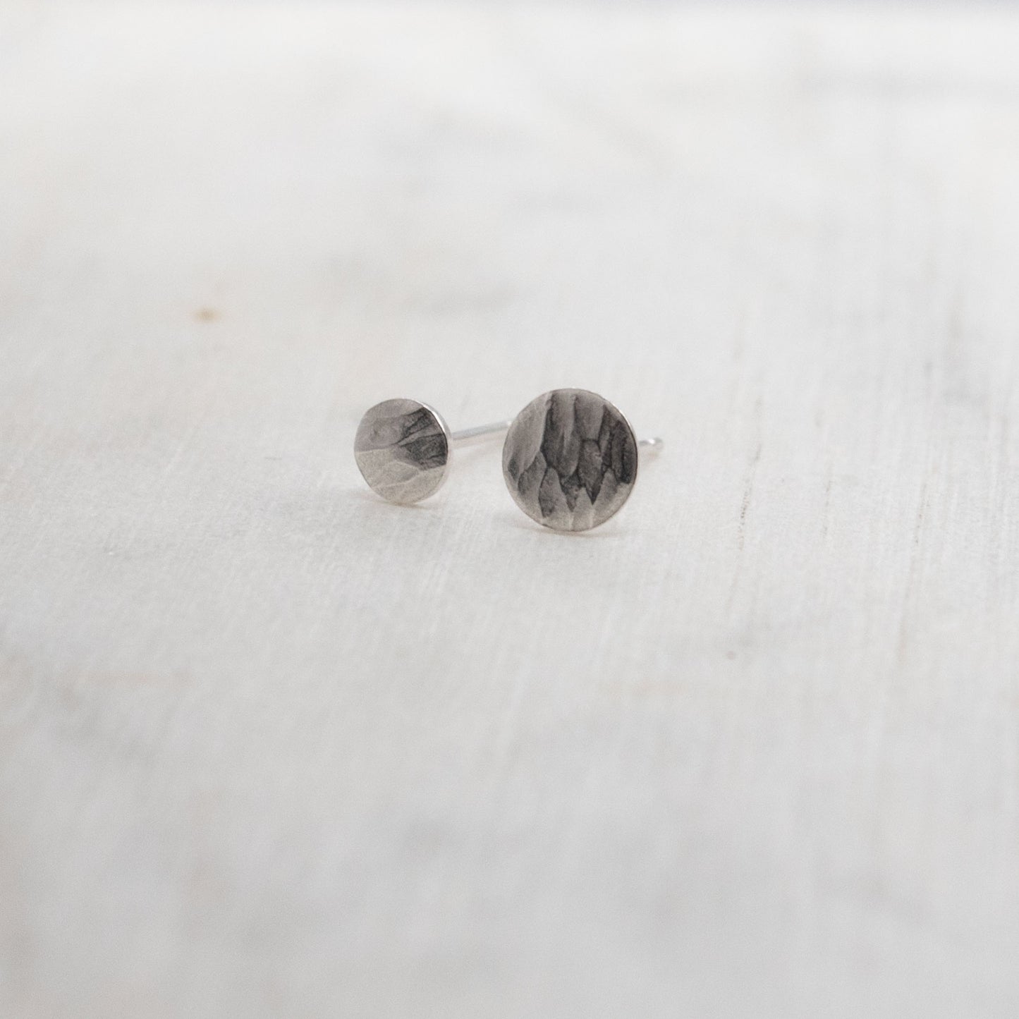 Boucles d'oreilles asymétriques en argent martelé posées sur une surface claire. Design minimaliste avec une texture organique et naturelle, idéales pour un usage quotidien.