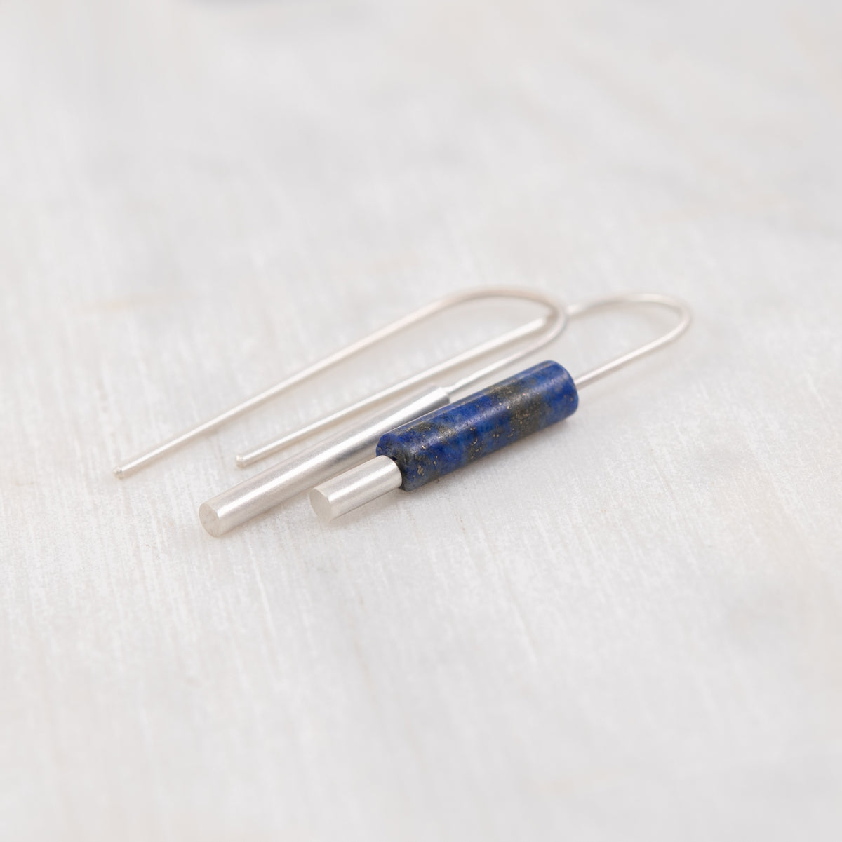 Une paire de boucles d’oreilles en fil d’argent ornées d’une petite perle cylindrique en lapis-lazuli bleu, présentée sur un fond blanc texturé.