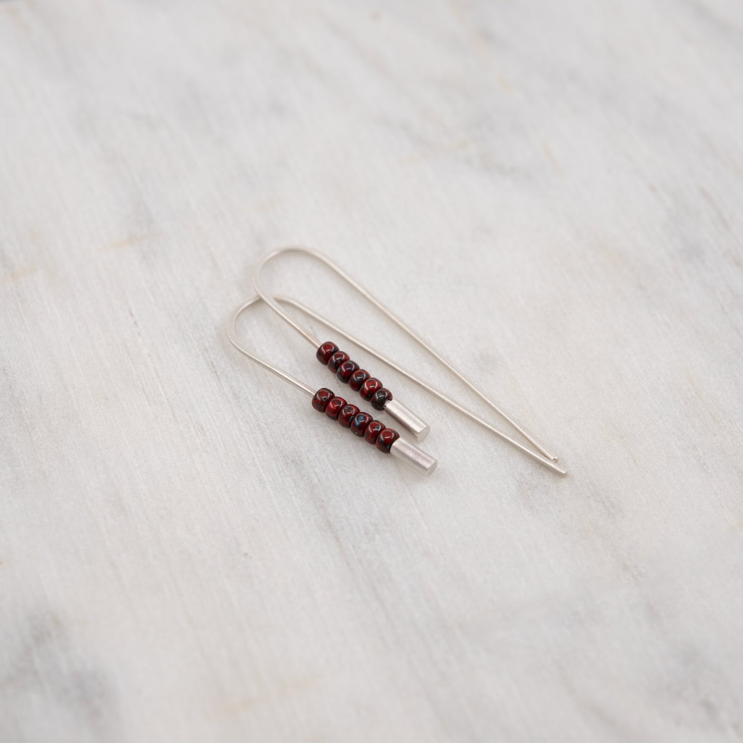Gros plan des boucles d’oreilles traversantes en argent massif brossé et perles rouges Miyuki, présentées sur une surface en marbre clair.