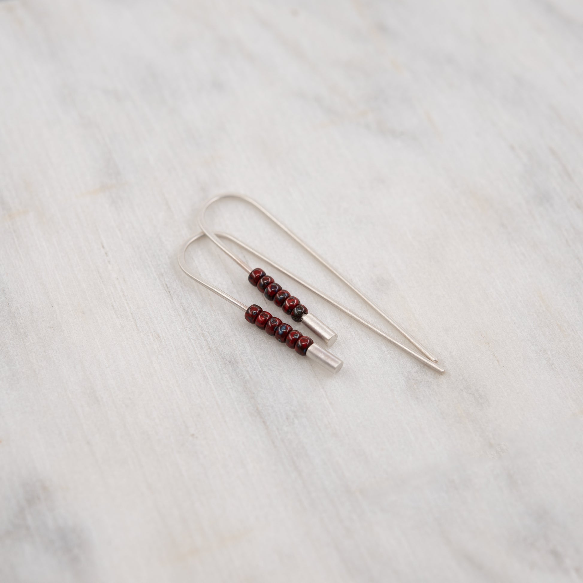 Gros plan des boucles d’oreilles traversantes en argent massif brossé et perles rouges Miyuki, présentées sur une surface en marbre clair.